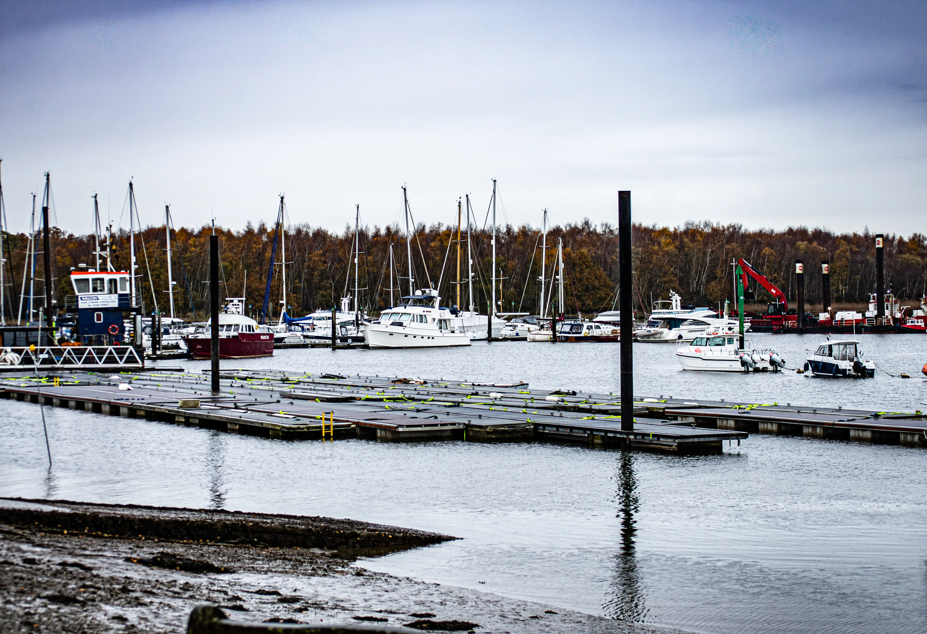 The image depicts a marina with various boats and yachts docked at the piers. The scene is set on a cloudy day with calm water, and the background features a forested area with autumn foliage. The marina is equipped with several docks and mooring lines, and there are a few buildings and structures visible, including a control tower and a flagpole with a red and green flag.