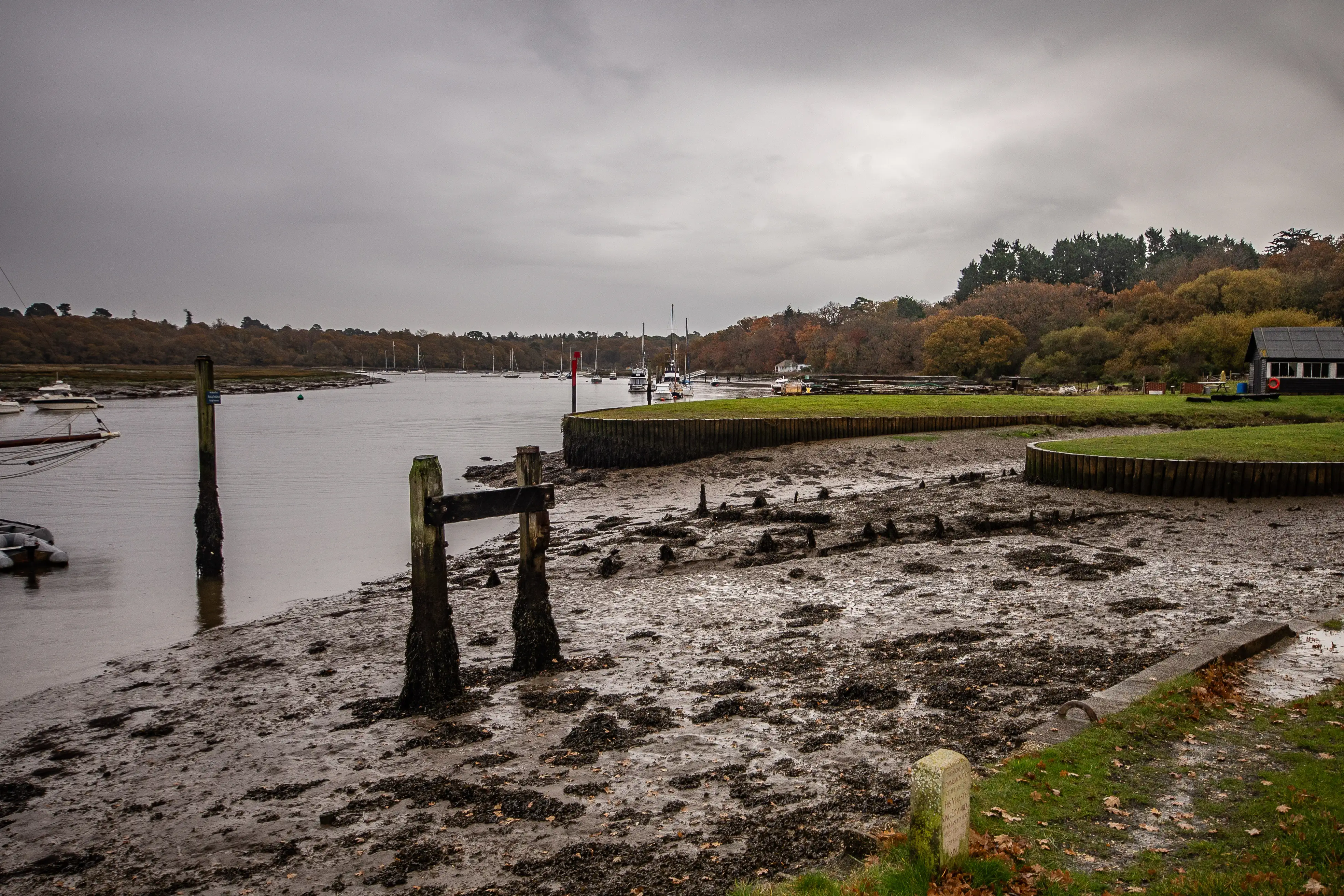 The image depicts a tranquil riverside scene with a muddy shore, wooden posts, and boats anchored in the water. The area is surrounded by trees with autumn foliage, and the sky is overcast, suggesting a calm, possibly rainy day.
