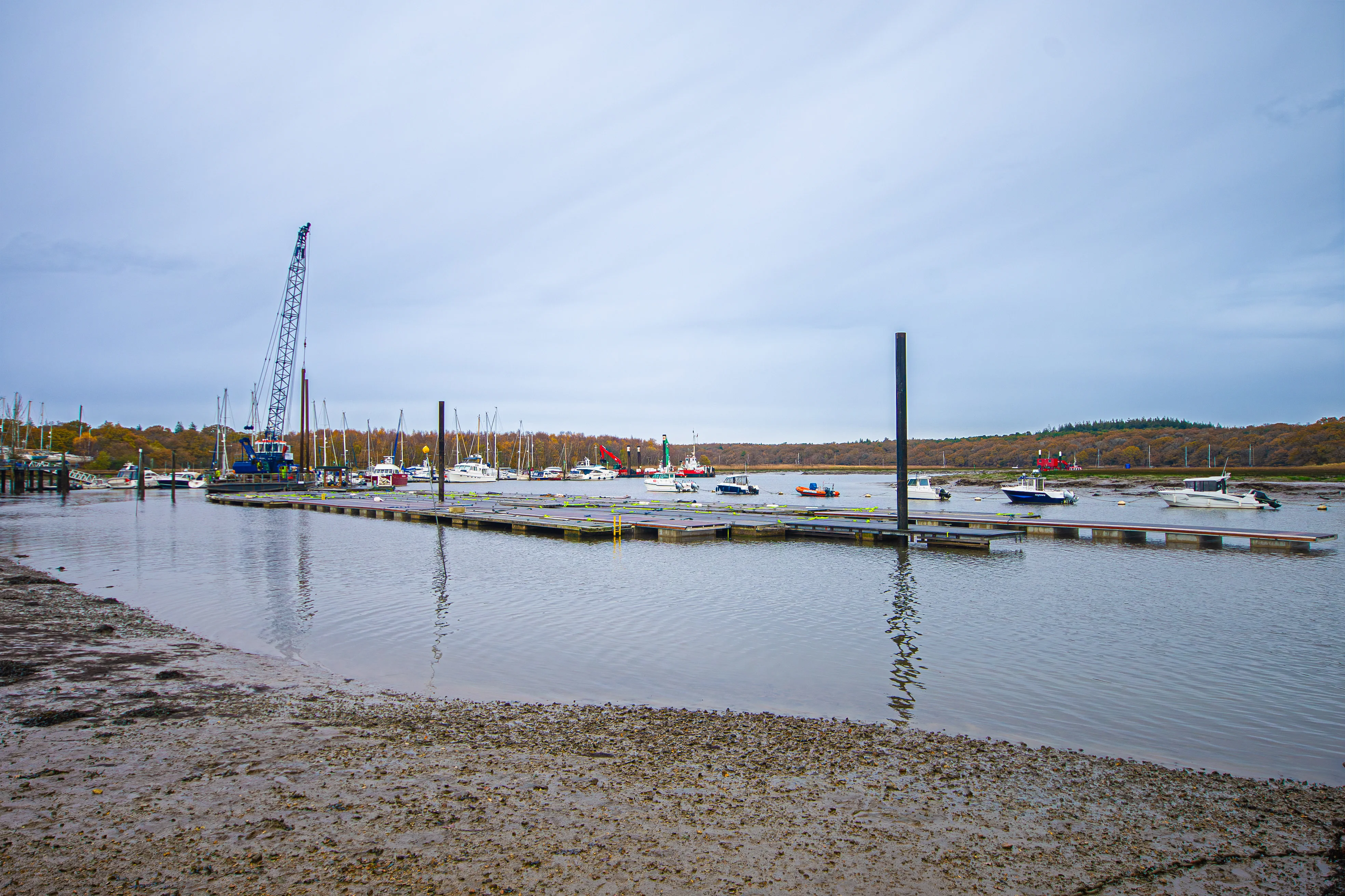 The image depicts a harbor scene with various boats and yachts docked at piers. There are cranes and other industrial equipment in the background, indicating ongoing construction or maintenance work. The water is calm, and the shoreline is visible in the foreground with some exposed seabed. The sky is overcast, suggesting it might be a cloudy day. The overall atmosphere is quiet and serene, typical of a harbor setting.