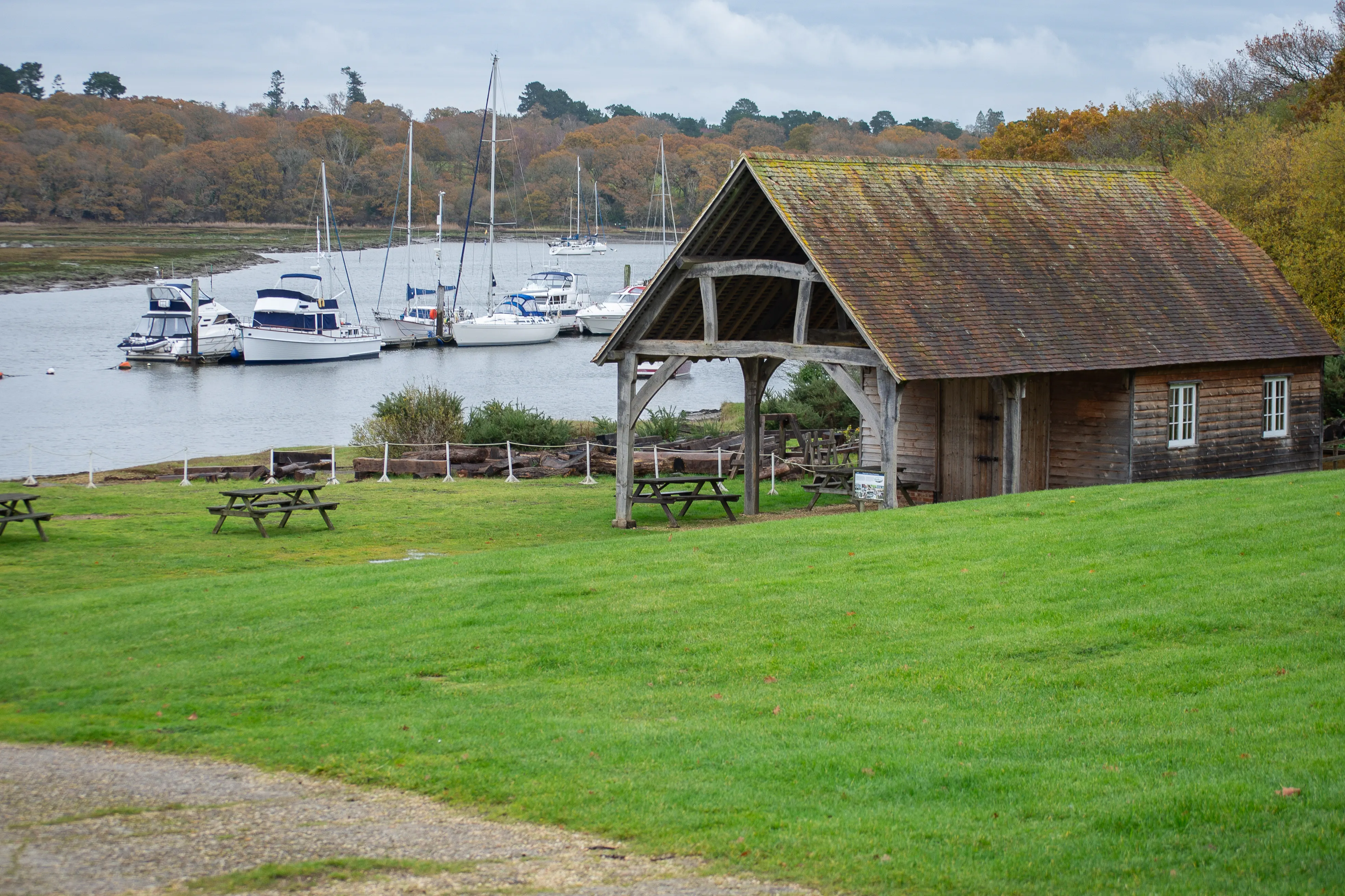The image depicts a serene waterfront scene with a rustic wooden structure and several boats docked in the background. The foreground features a grassy area with picnic tables and a small shelter. The overall atmosphere is peaceful and inviting, suggesting a tranquil spot for relaxation and outdoor activities.