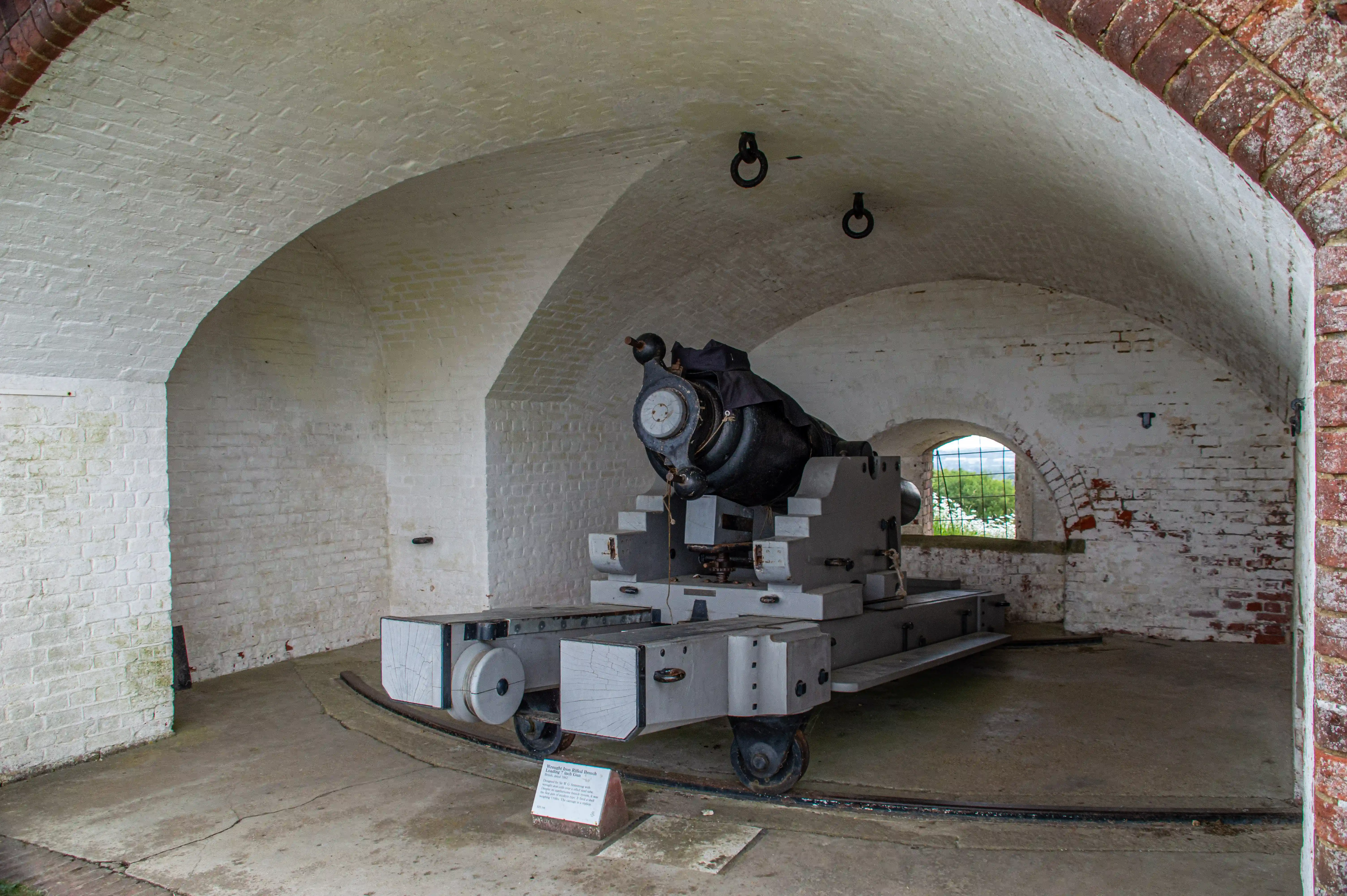 The image depicts an old cannon mounted on a wheeled carriage inside a brick-lined fortification. The fort has arched ceilings and appears to be part of a historical military site. The cannon is positioned to fire through an arched opening in the wall, with a small informational plaque in front of it.