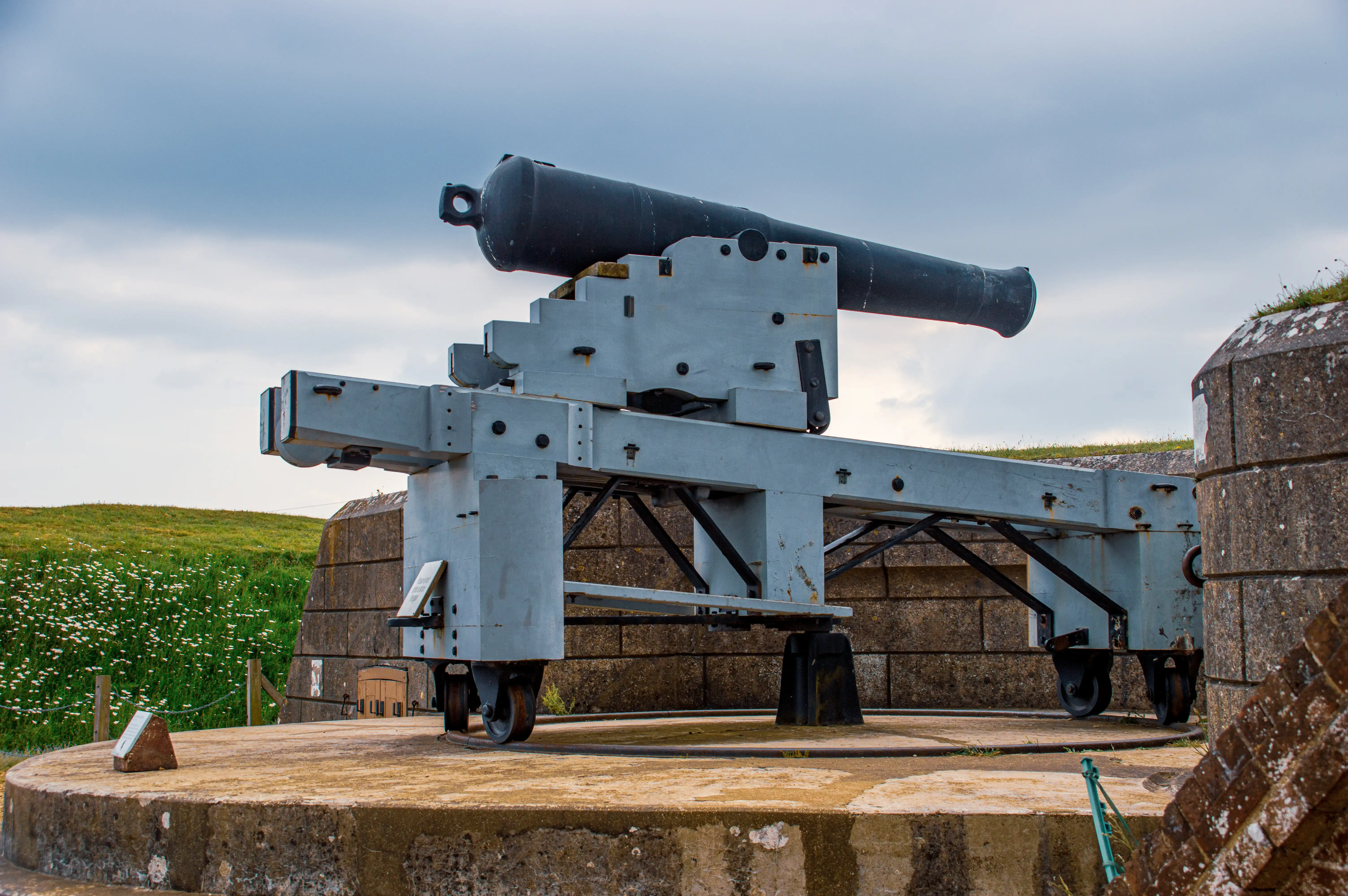 The image depicts an old, large-caliber artillery gun mounted on a mobile carriage, situated on a concrete platform. The gun is positioned outdoors, with a grassy hill and a partly cloudy sky in the background. The structure supporting the gun appears to be made of metal and is designed to allow the gun to be moved and aimed in different directions. The surrounding area includes concrete fortifications, suggesting a historical military installation or fort.