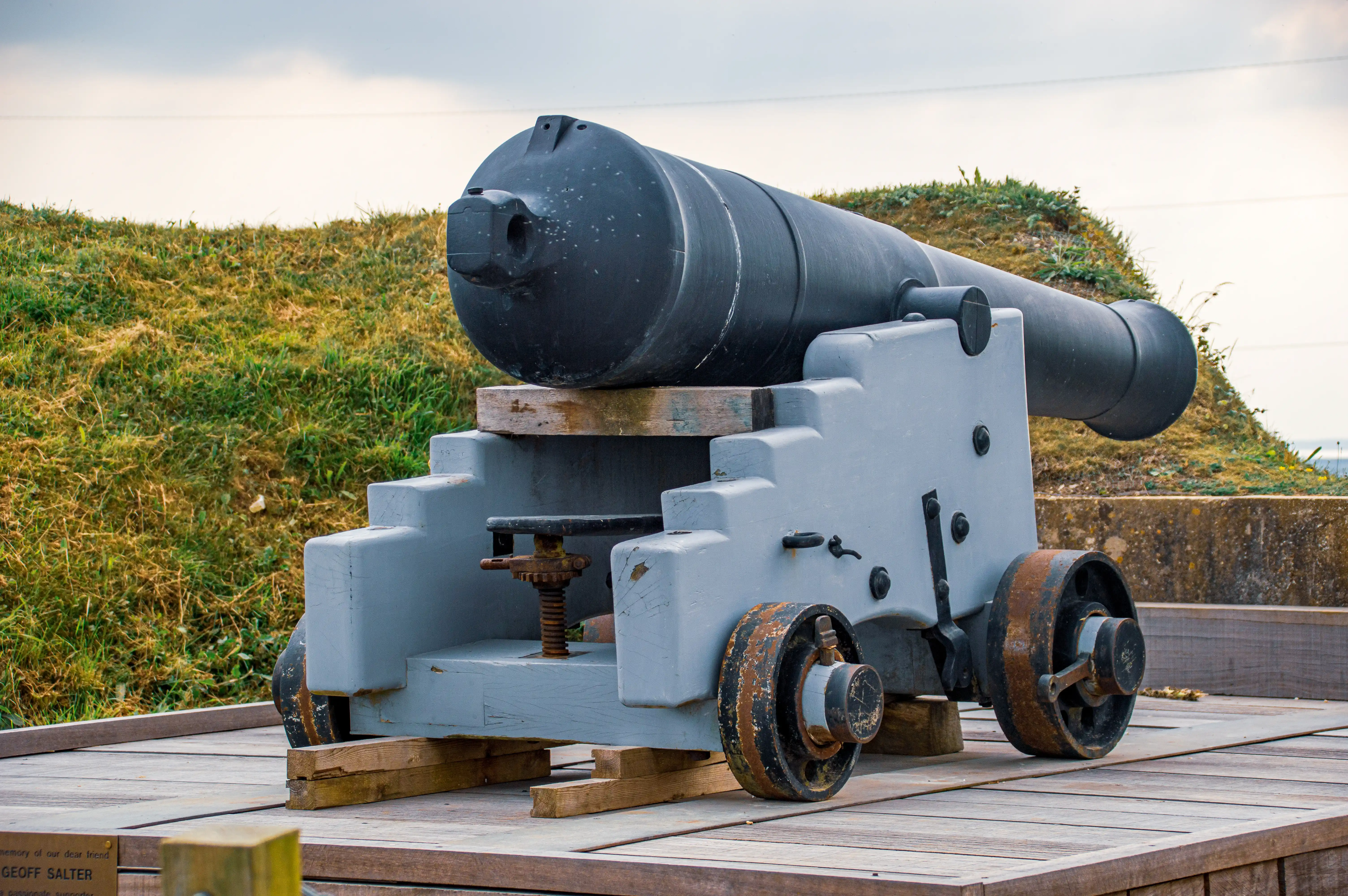 The image depicts an old cannon mounted on a wooden carriage, positioned on a wooden platform. The cannon is black with a cylindrical barrel and is aimed slightly upward. The carriage supporting the cannon is gray with rusted metal wheels and various mechanical components. The background shows a grassy hill with some vegetation and a partly cloudy sky.