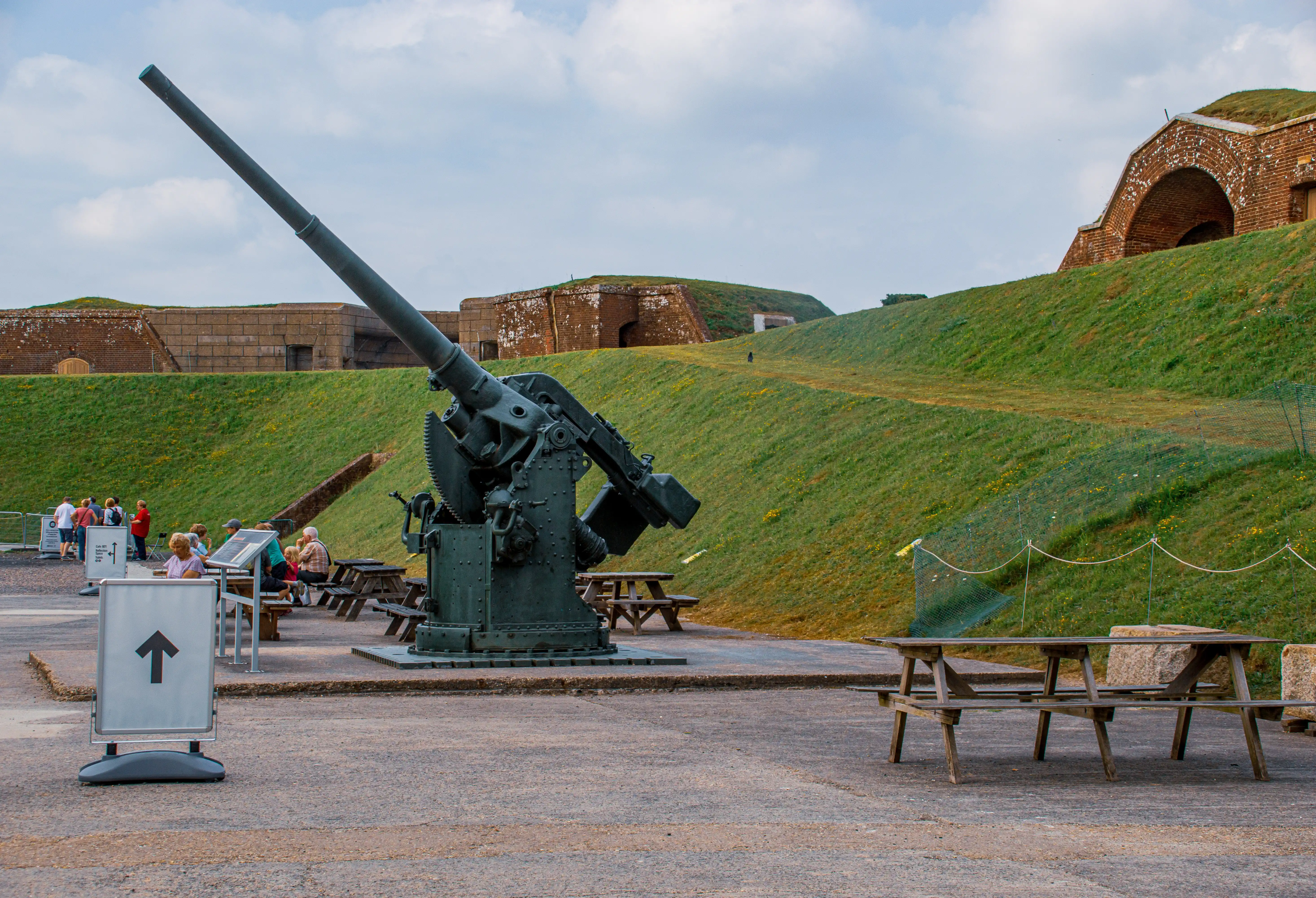 The image depicts an outdoor historical site featuring an old artillery gun on display. The gun is mounted on a concrete platform, with informational signs and benches around it. In the background, there are grassy hills with brick structures and arches. A few people are seen reading the informational signs and sitting on the benches. The sky is partly cloudy, suggesting a calm day.