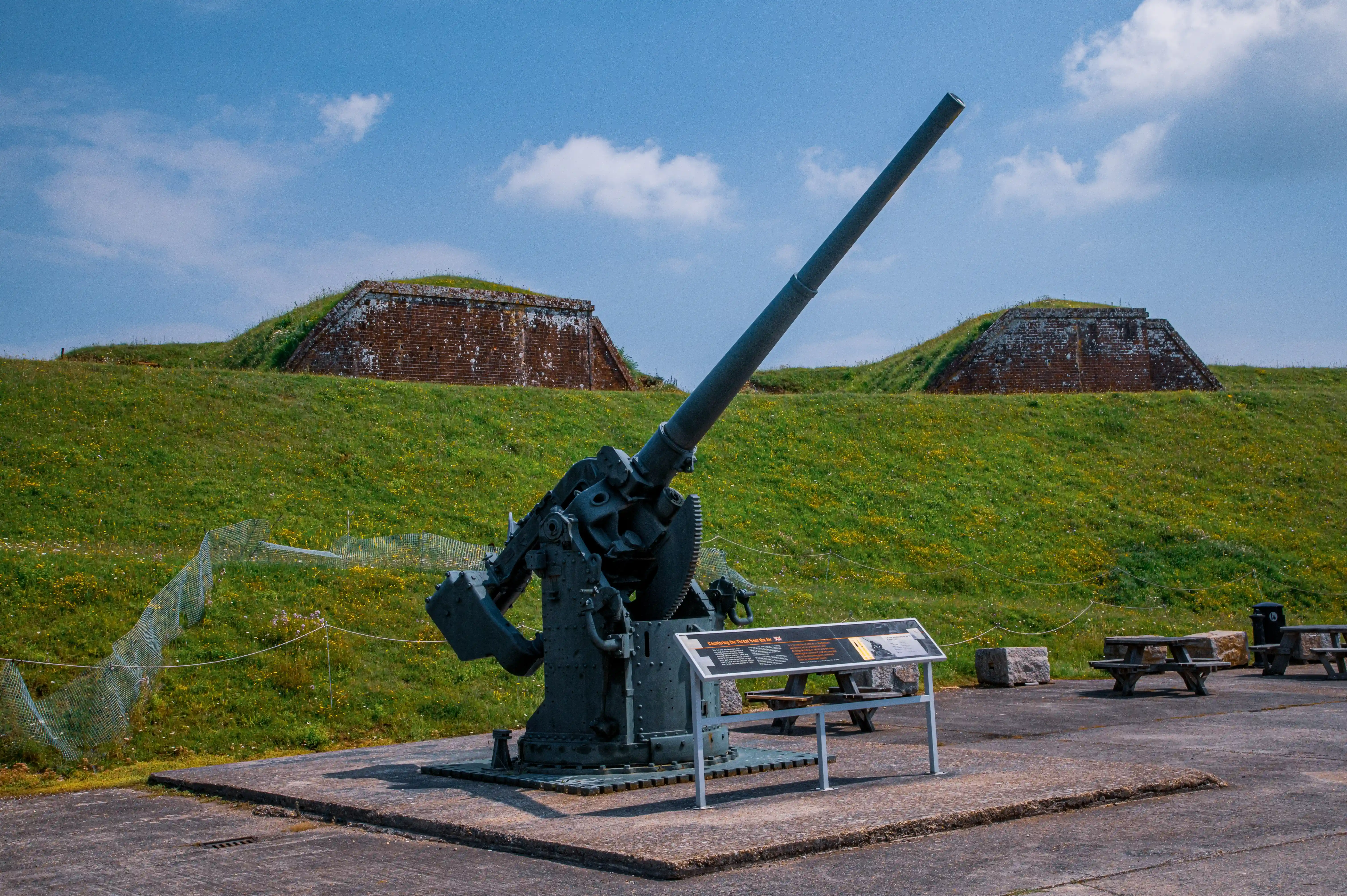 The image shows an outdoor historical site featuring a large, mounted artillery gun on display. The gun is positioned on a concrete platform with informational plaques nearby. In the background, there are two grassy mounds with brick structures on top, likely part of the fortification. The area is fenced off with metal mesh and rope barriers, and there are picnic tables and a trash can in the vicinity, indicating it is a public, possibly educational, site.
