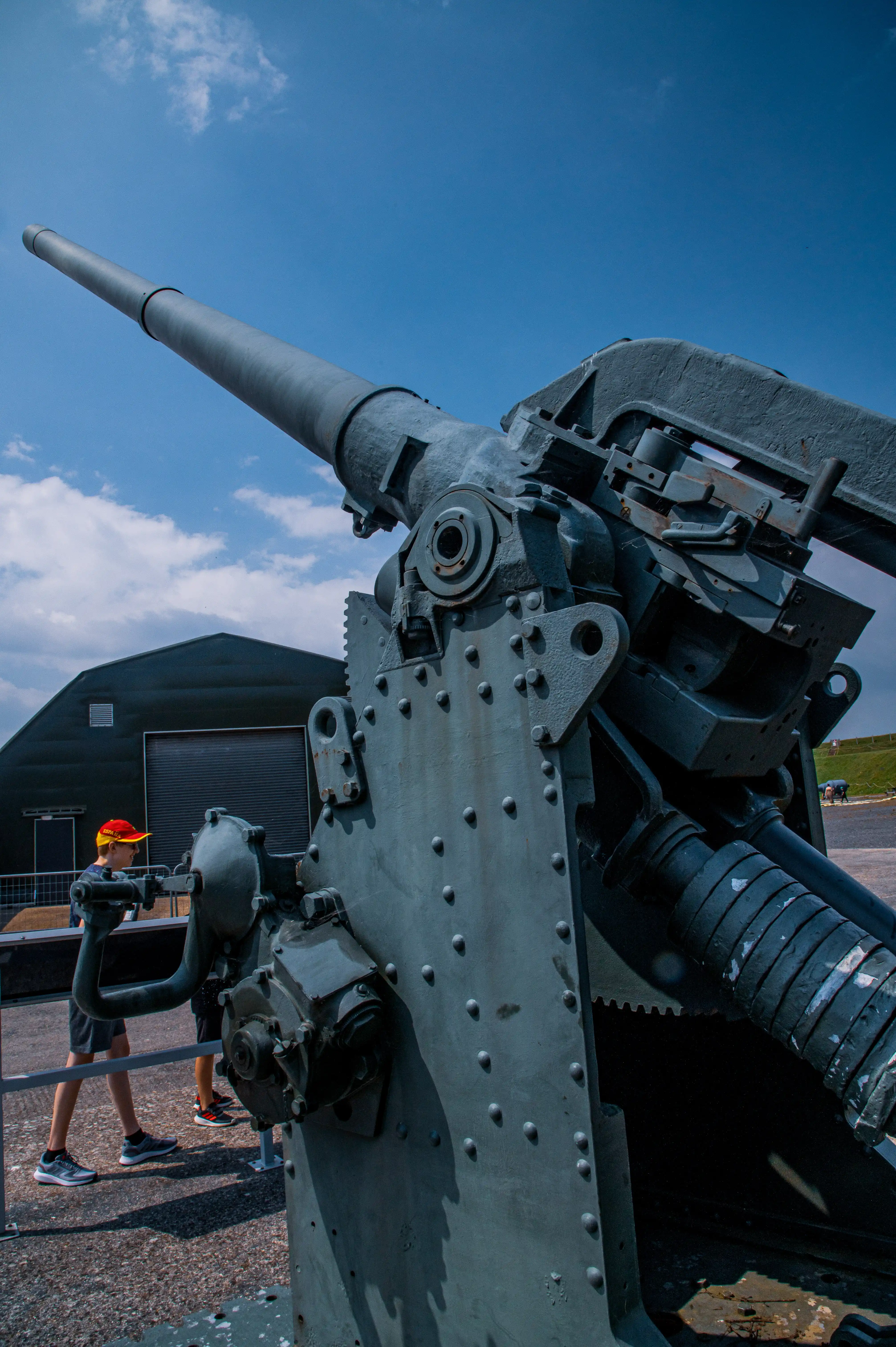 The image shows a large, detailed view of a historical artillery piece, likely from a museum or historical site. The cannon is prominently displayed with intricate mechanical details visible. In the background, there is a person wearing a red cap and dark clothing, standing near a barrier and observing the cannon. The sky is clear with a few clouds, and there is a building with a large, closed rolling door behind the person. The overall scene suggests an outdoor exhibit where visitors can view and learn about historical military equipment