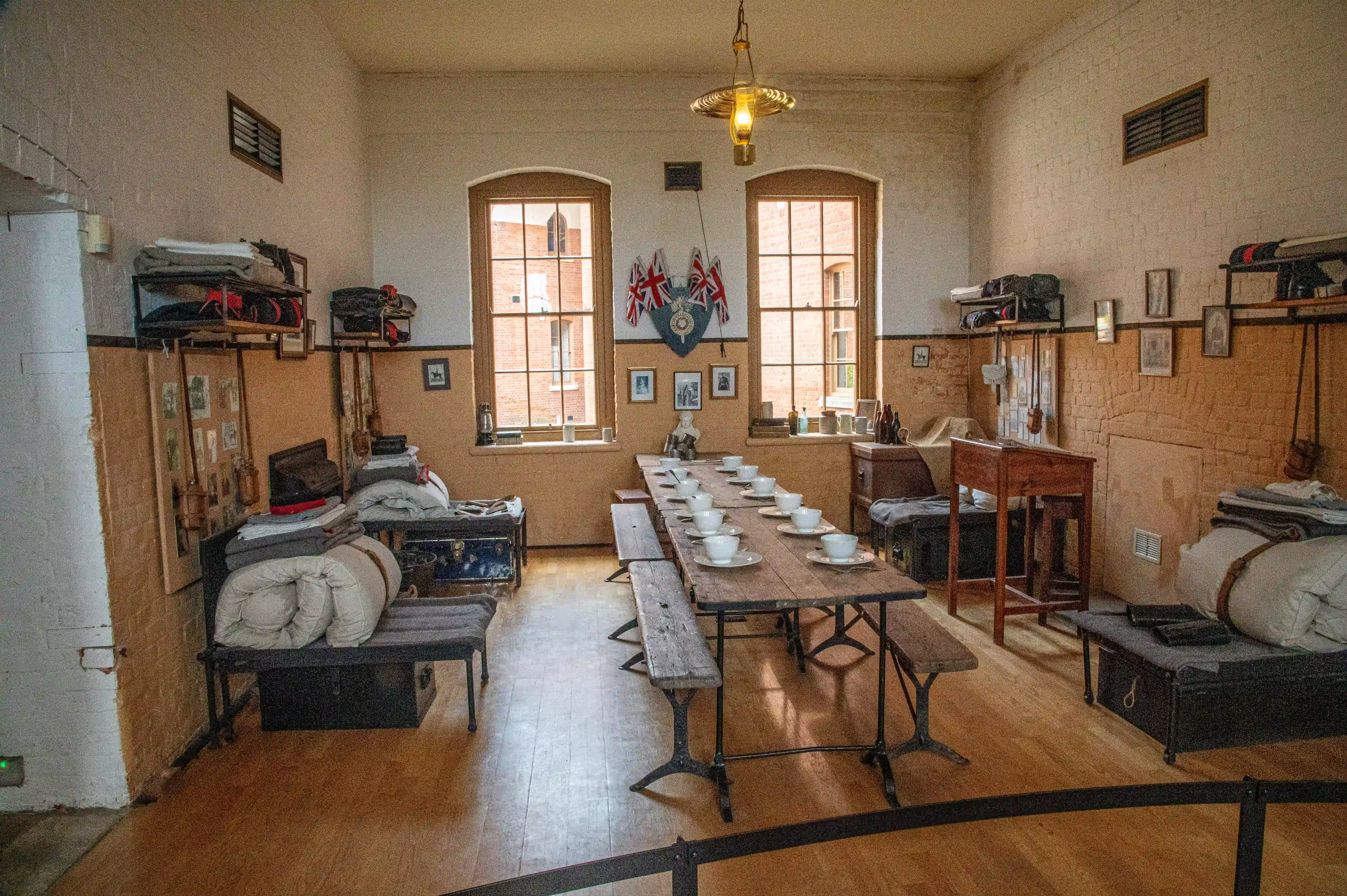 The image depicts a historical room with bunk beds lined along the walls, a central wooden table set with bowls and plates, and various personal items and decorations. The room features two large windows with natural light, a shield with flags above them, and shelves holding more personal belongings. The overall setting suggests a communal living space, possibly from a military or institutional context, with a focus on simplicity and functionality.