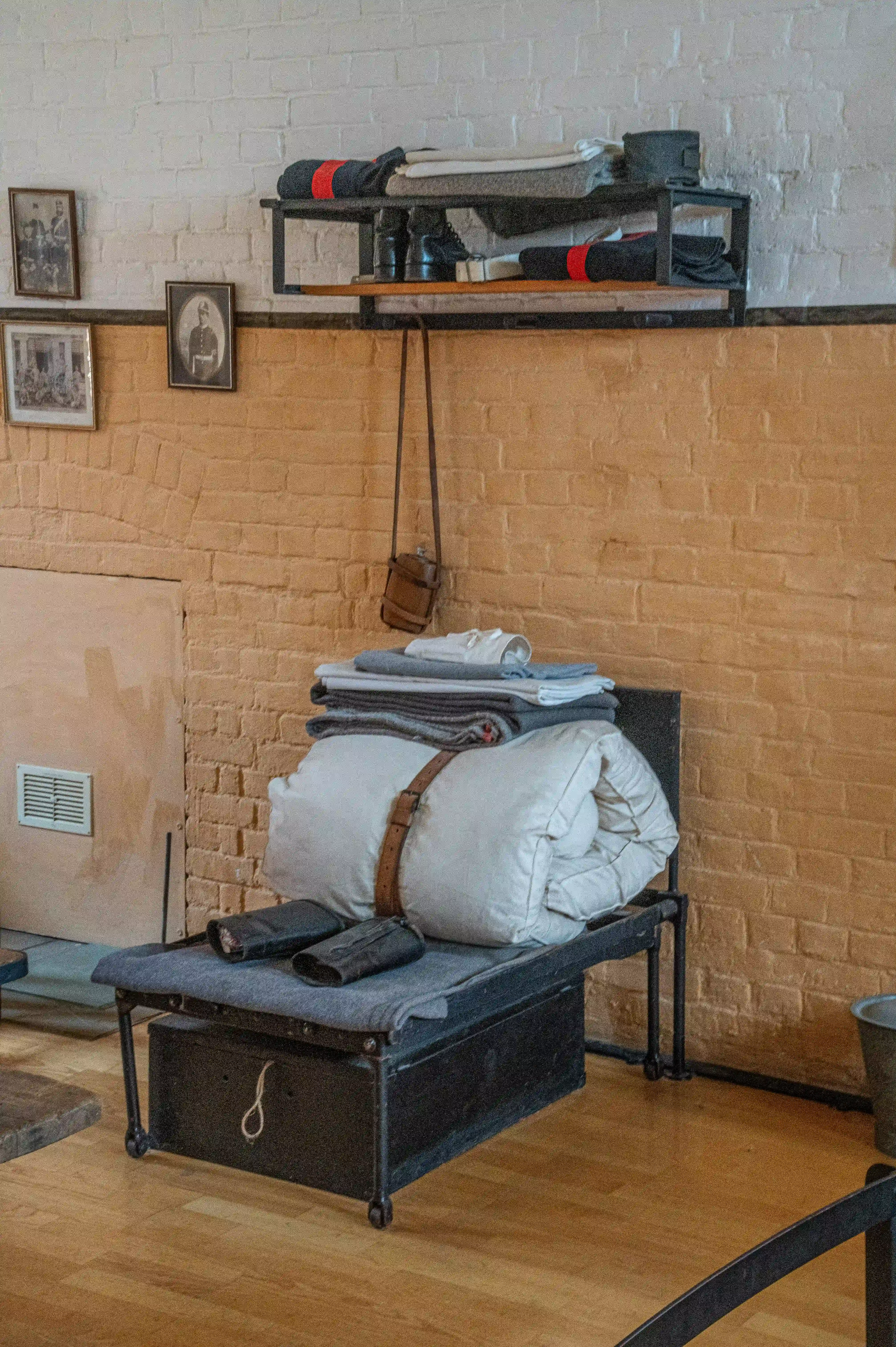 The image depicts a rustic, industrial-style room with a metal-framed bed or couch that has a grey cushion and a large white pillow. Above it, there is a metal shelf holding various items, including folded clothes, shoes, and a small box. The walls are made of exposed brick, and there are several black-and-white photographs or portraits hanging on the wall. A small brown bag hangs from a hook on the wall, and there is a vent visible on the left side of the image. The floor is made of wooden planks.