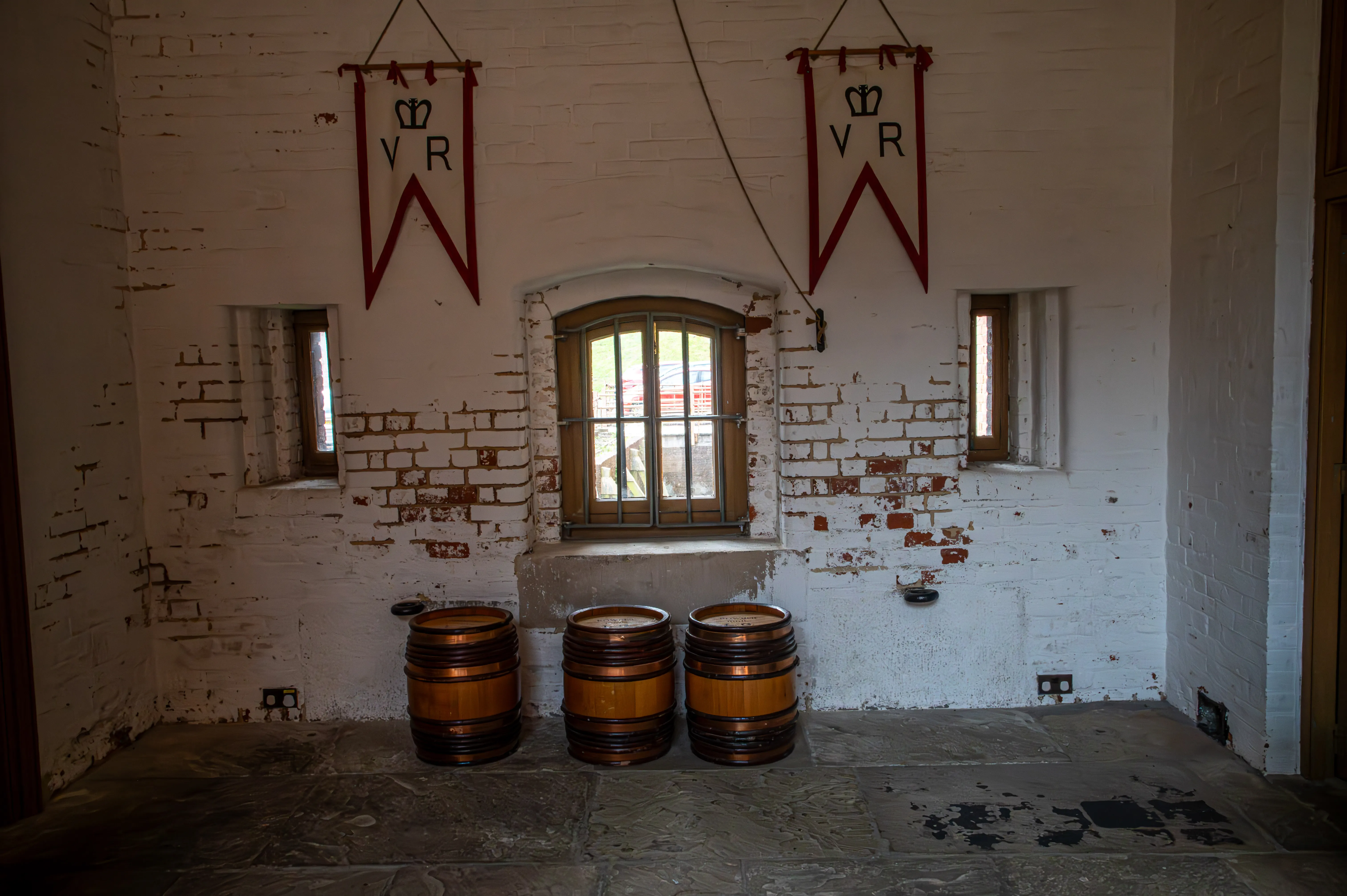 The image depicts an interior room with three wooden barrels placed on a stone floor. The room has white brick walls with visible signs of wear and aging. There are three windows, one of which is larger and arched, allowing natural light to enter. Two red banners with the letters 'VR' and a crown symbol hang from the ceiling.