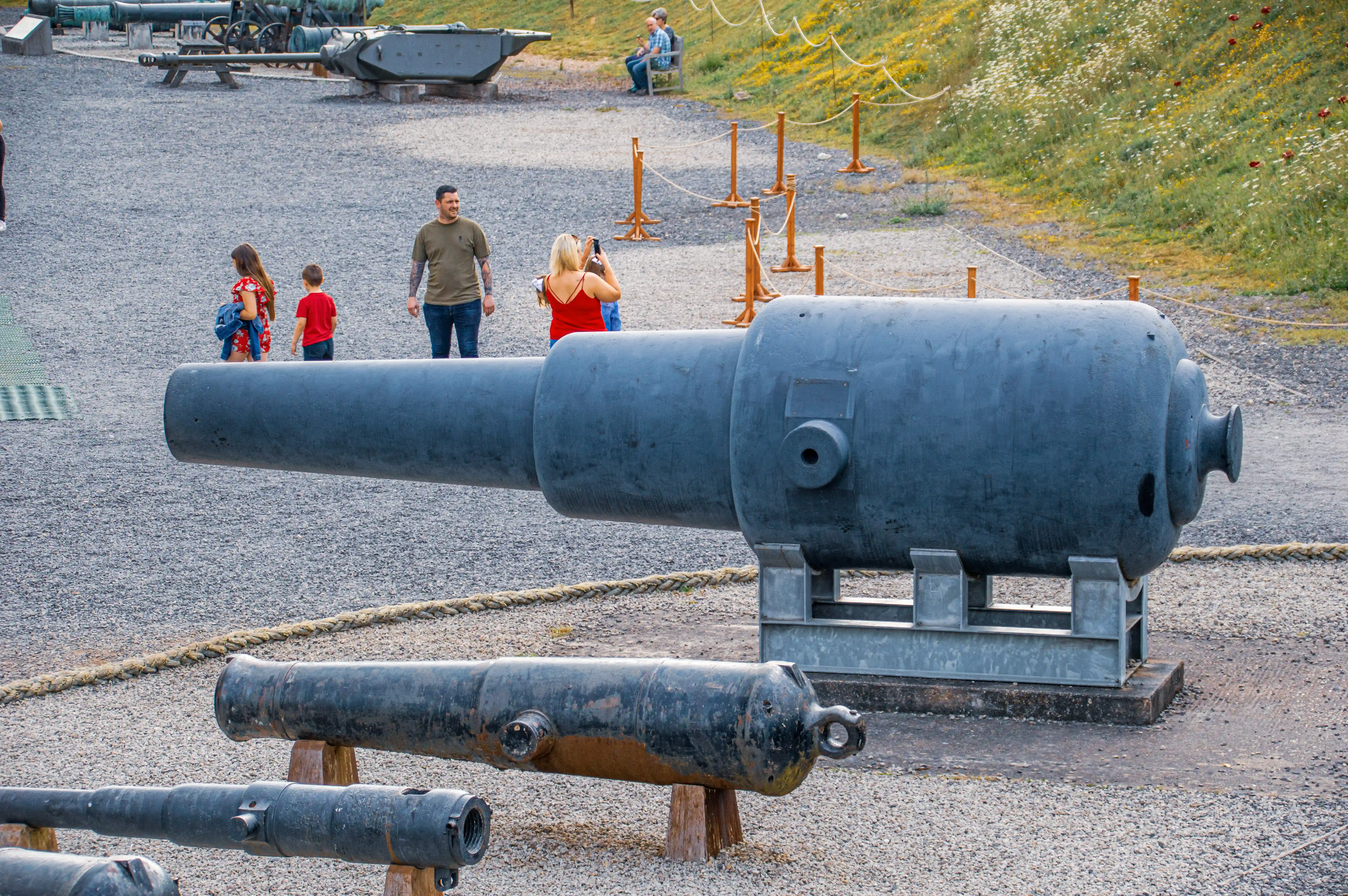 The image shows a historical site with several large cannons on display. Visitors are seen walking around and taking photos. The area is surrounded by a rope barrier and has a gravel surface with some greenery in the background.
