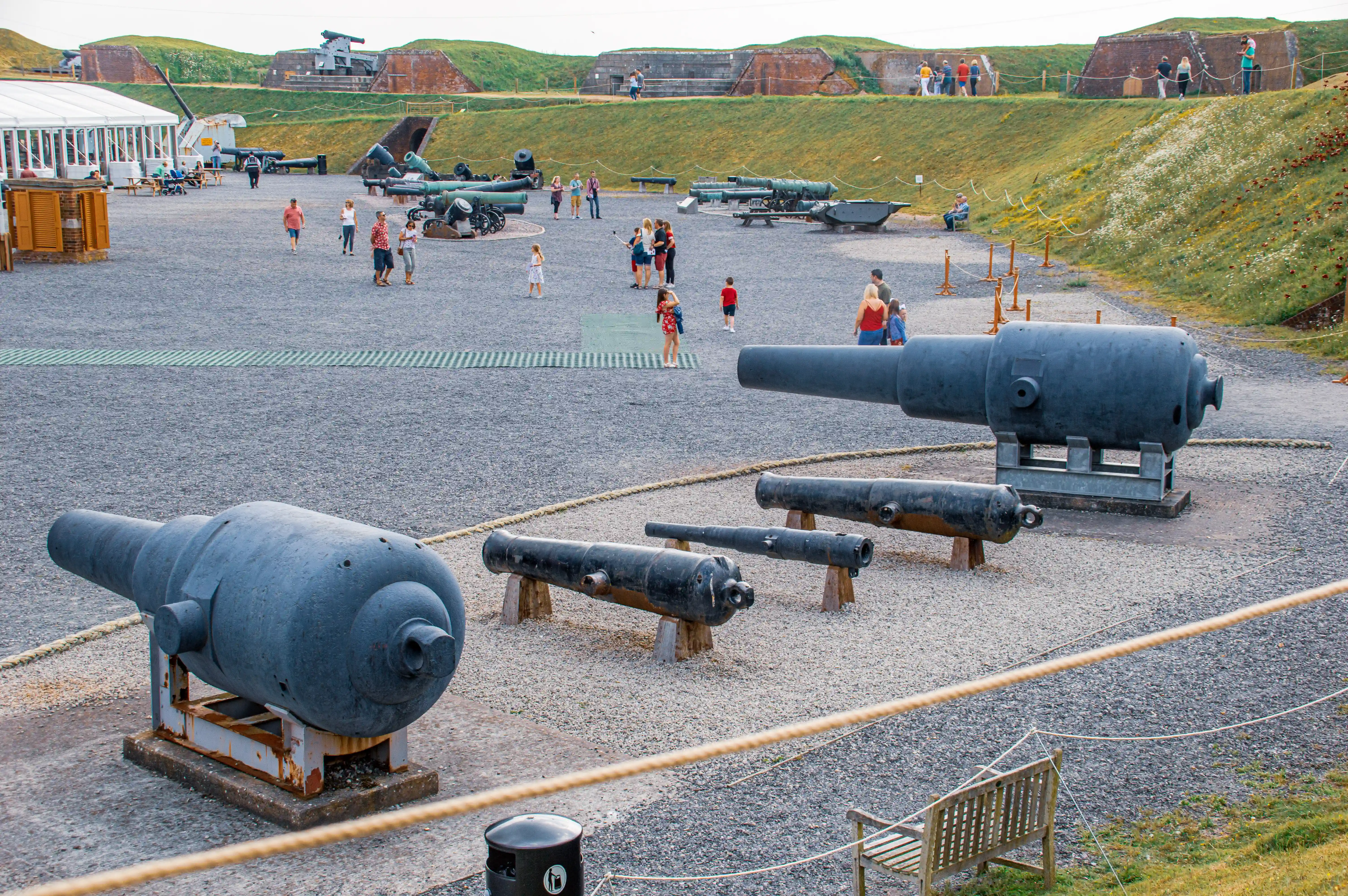 The image depicts an outdoor historical site with various large cannons and artillery pieces on display. The area is surrounded by grassy embankments and features several groups of people walking around and observing the exhibits. There is a tent-like structure on the left side, possibly serving as an information or ticket booth. The ground is gravel, and the cannons are arranged in an open space, suggesting a museum or historical fort setting.