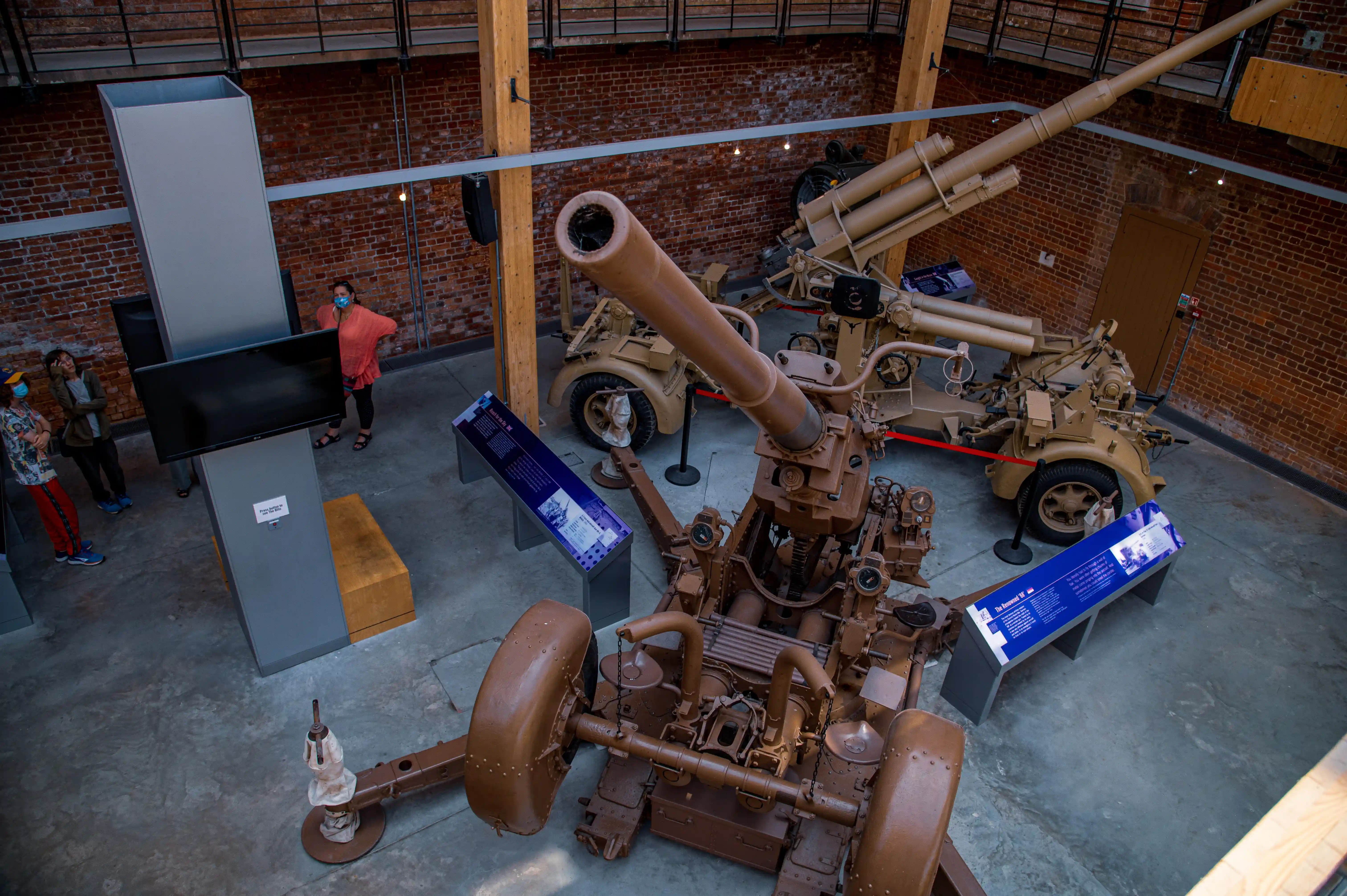 The image shows a museum exhibit featuring a large, antique artillery piece mounted on a wheeled carriage. The exhibit is housed in a spacious, brick-walled room with high ceilings and large windows. Several informational plaques are placed around the artillery piece, providing details about its history and specifications. Two people are present in the room, observing the exhibit. The overall atmosphere suggests a historical or military museum setting.