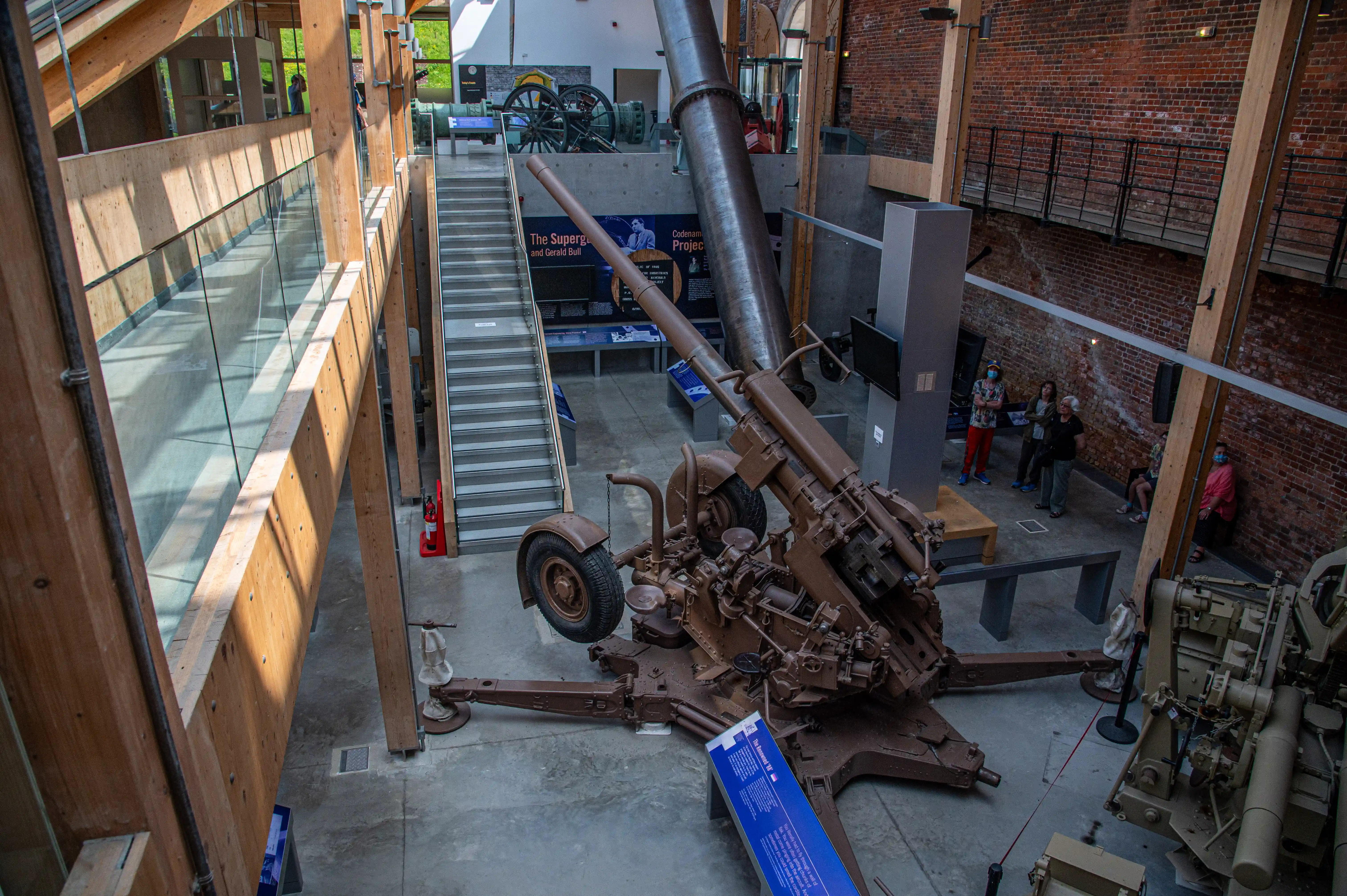 The image depicts an indoor museum space featuring large artillery pieces on display. The museum has a modern design with wooden and glass elements, and brick walls. Visitors are seen observing the exhibits, and there are informational placards near the artifacts. The space is well-lit with natural light, and there is a staircase in the center of the image.
