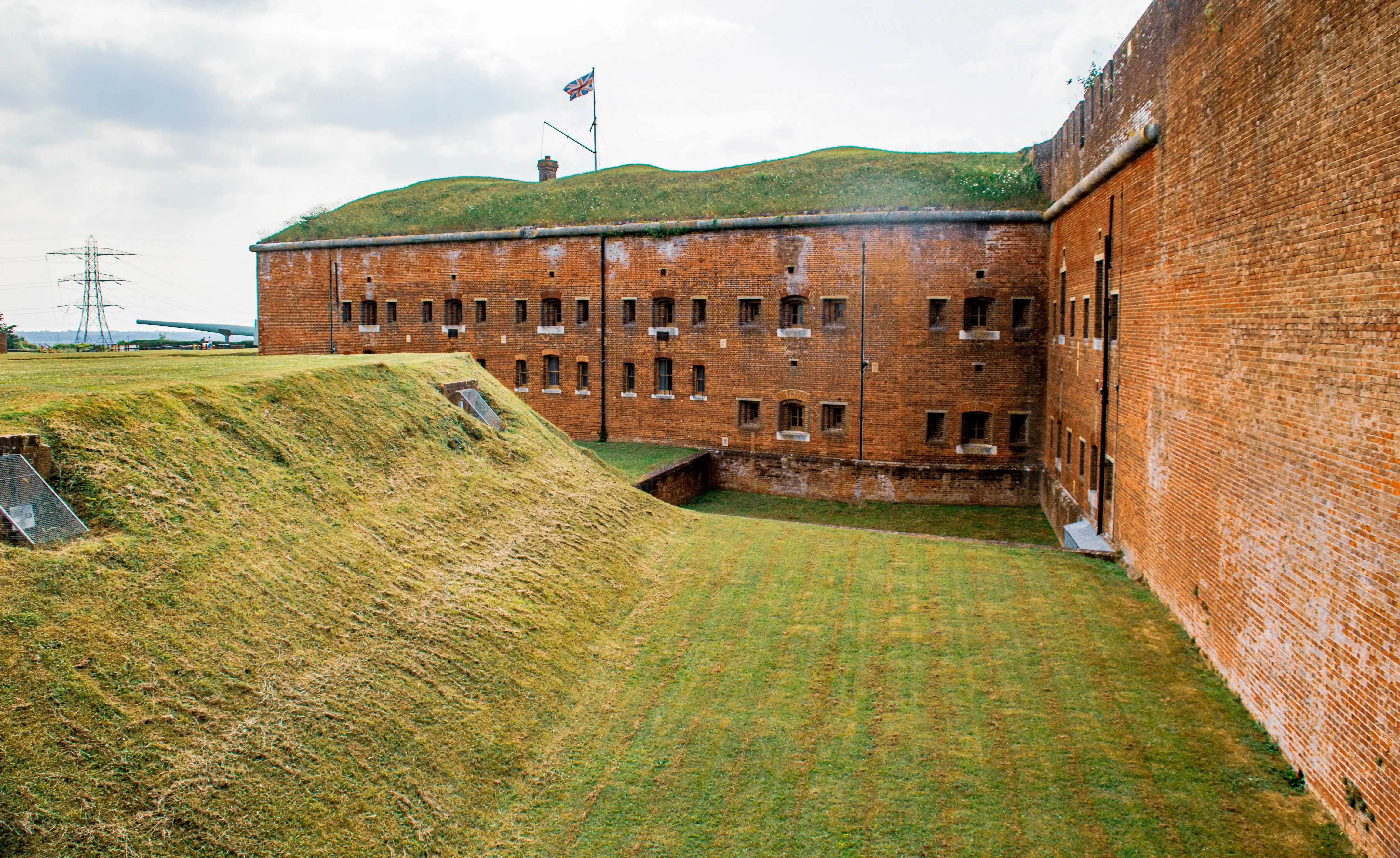 The image depicts a historic fortification with red brick walls and a grass-covered roof. The structure has multiple small windows and is surrounded by a dry moat. A flagpole with a flag flies atop the fort. The fort is situated in an open area with some modern infrastructure visible in the background.