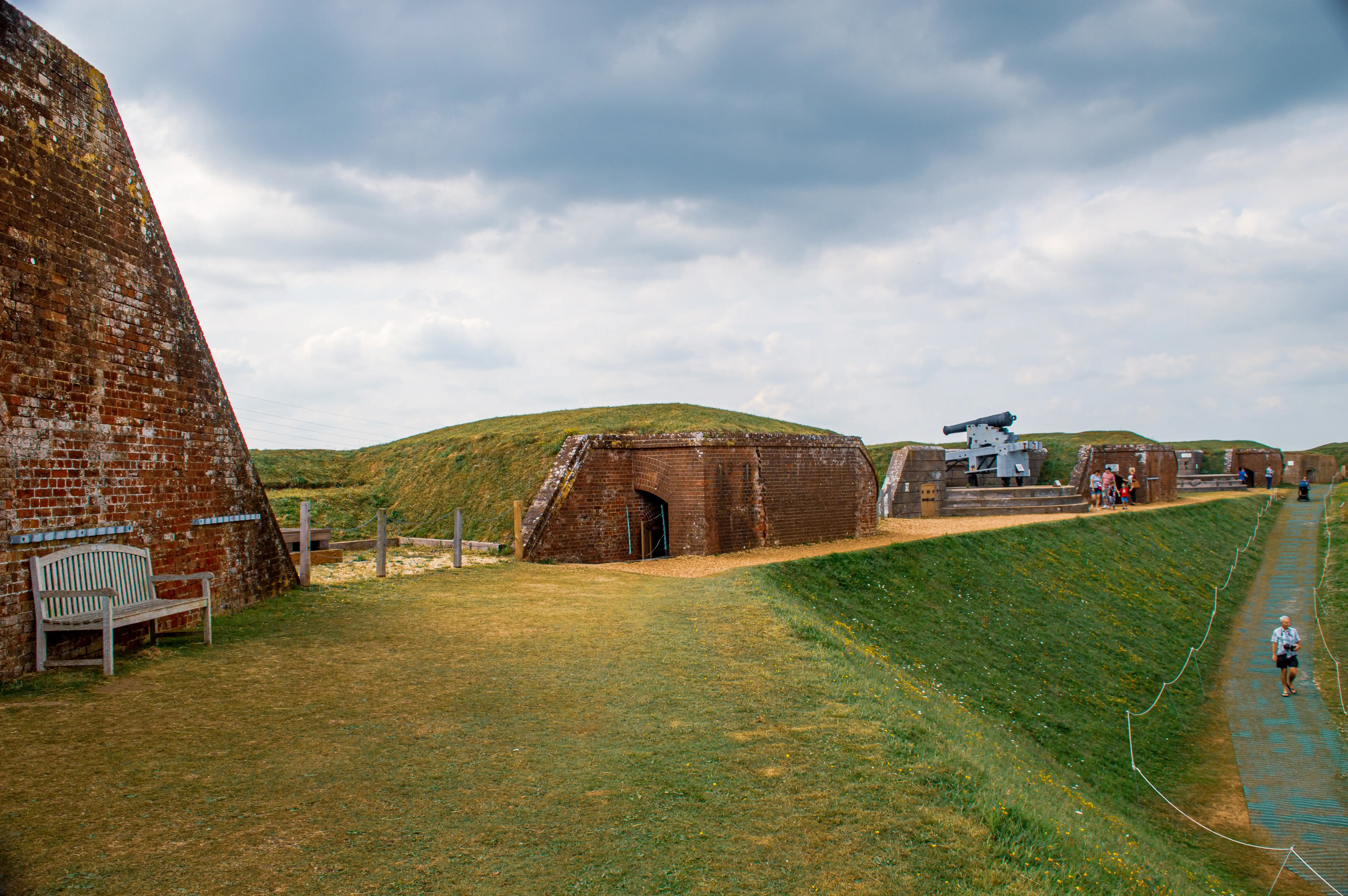 The image depicts a historical fortification with brick structures and grassy mounds. There are several cannons mounted on the mounds, and a few visitors are exploring the site. A bench is positioned near the brick wall on the left side of the image