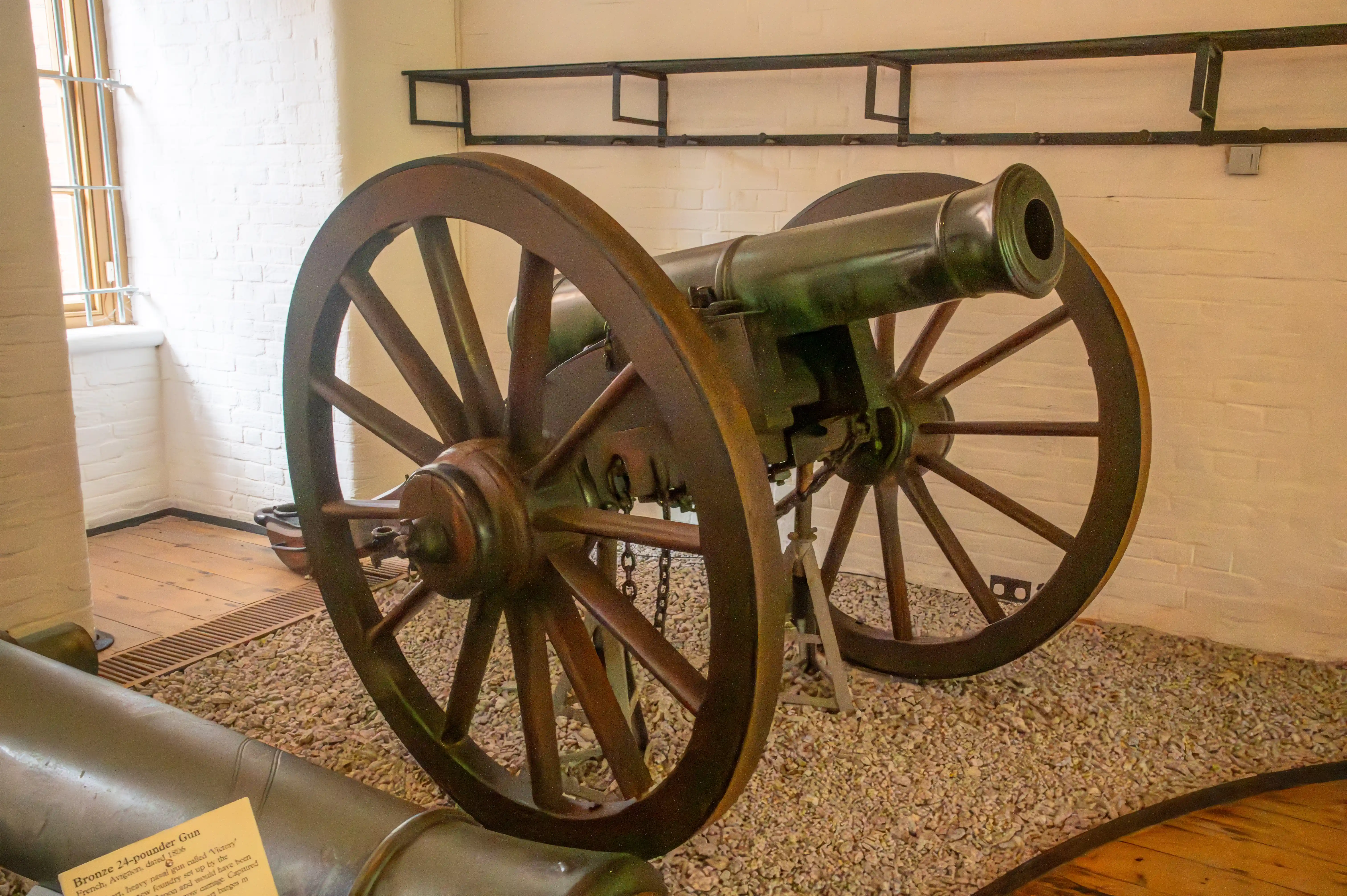 The image depicts an old cannon displayed in a museum-like setting. The cannon is mounted on large wooden wheels and is positioned on a bed of gravel. The surroundings include a white brick wall and a wooden floor, with another cannon partially visible in the foreground. There is a small informational plaque next to the cannon, providing details about it.