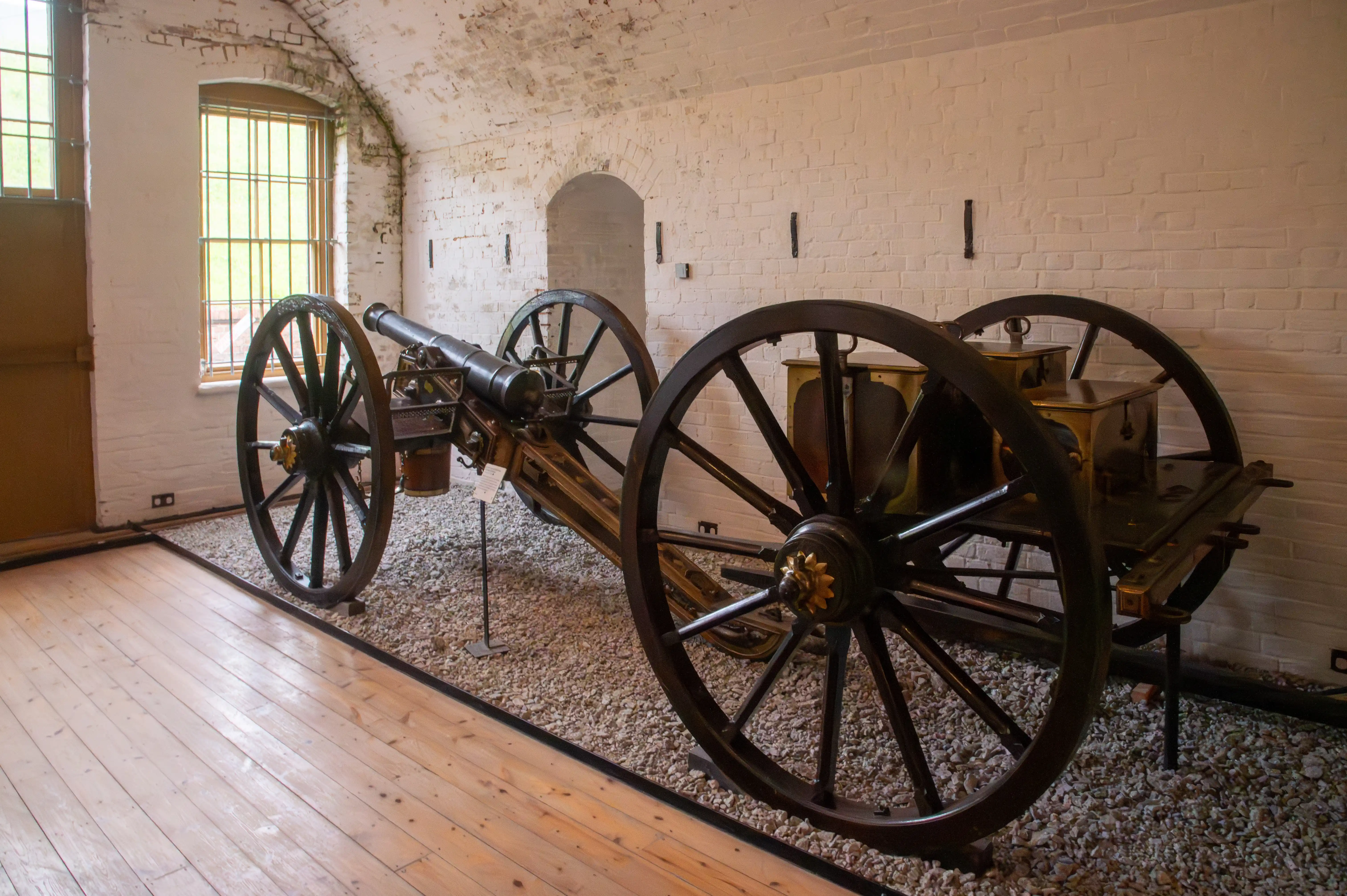 The image depicts two historical cannons displayed indoors on a gravel-covered floor with large wooden wheels. The room has white brick walls and several arched windows with metal bars. The setting appears to be a museum or historical exhibit.