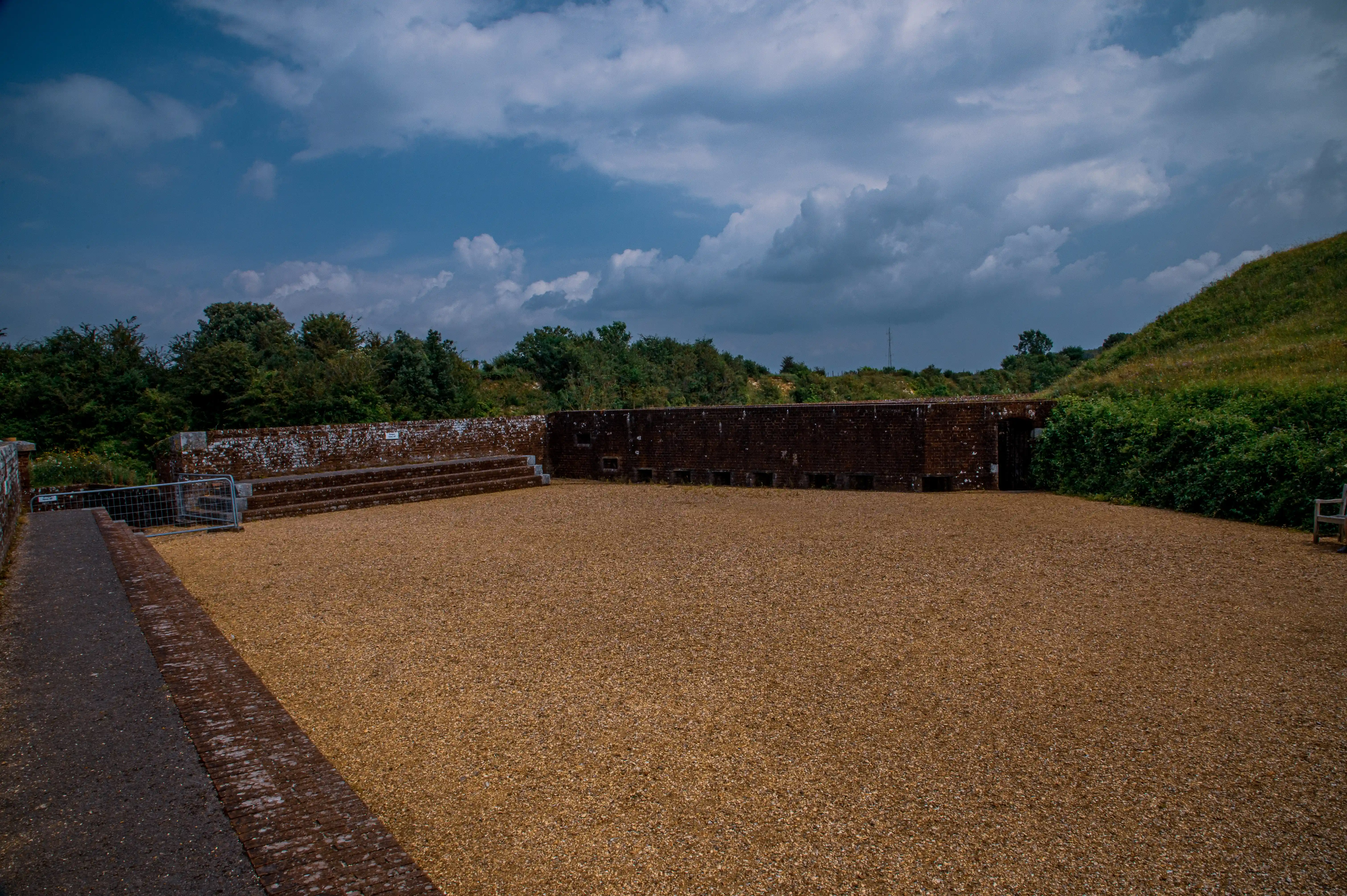 The image depicts an open, rectangular area with gravel ground, surrounded by a low brick wall and dense greenery. The sky above is partly cloudy, suggesting a mix of sun and clouds in the weather. The area appears to be a historical site or a ruin, possibly an old fort or amphitheater, with pathways leading into and around the space.