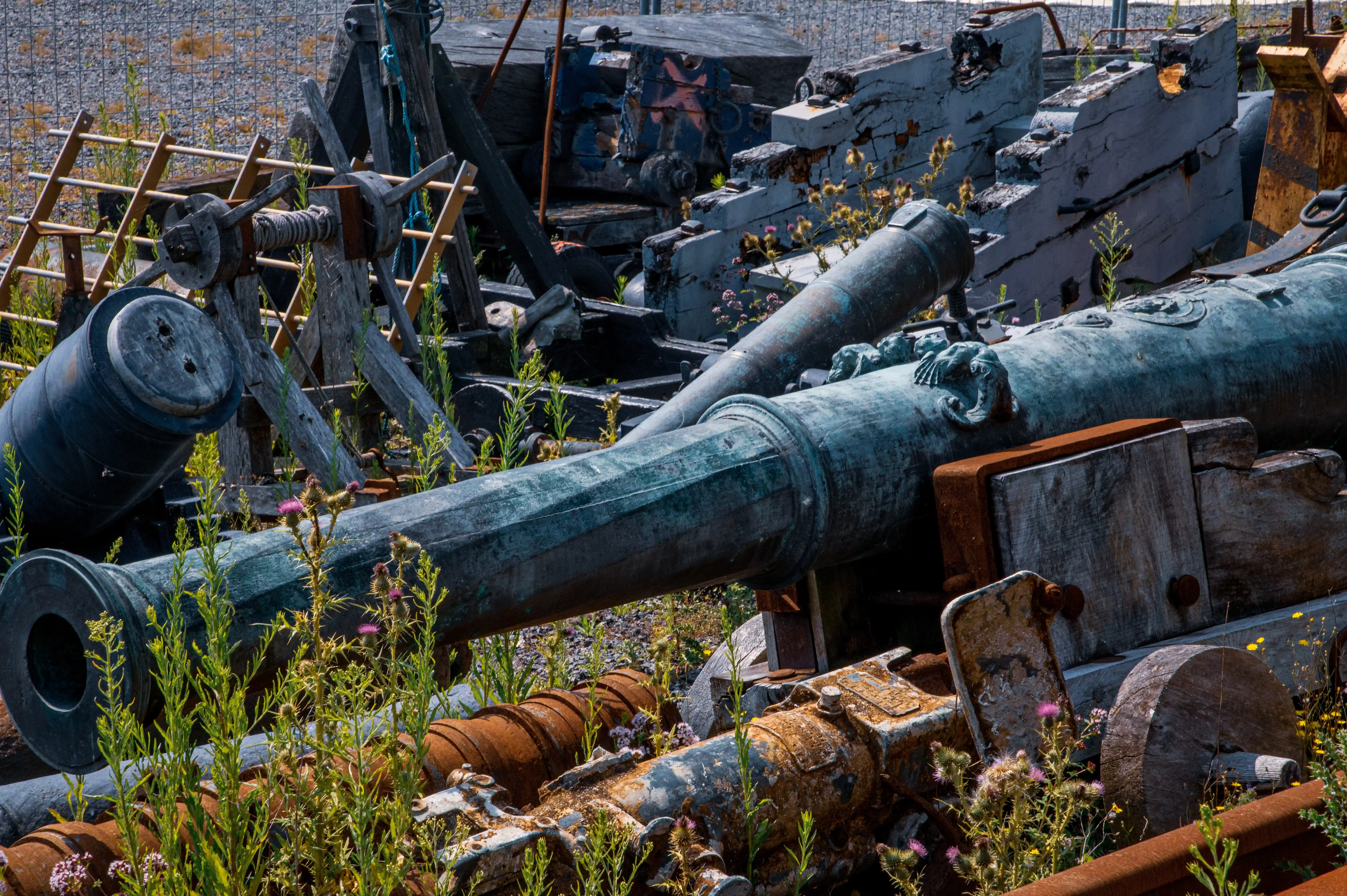 The image depicts an array of old, rustic cannons and various pieces of historical military equipment, likely from a bygone era. The cannons are weathered and covered in patina, with some adorned with decorative elements. The scene is set outdoors, with vegetation and wildflowers growing around and even through some of the equipment, indicating a state of disuse and abandonment. The background includes a stone wall and other rustic wooden structures, enhancing the historical ambiance. The overall atmosphere suggests a relic of historical warfare, possibly from a fort or a battlefield, now left to the elements and nature.