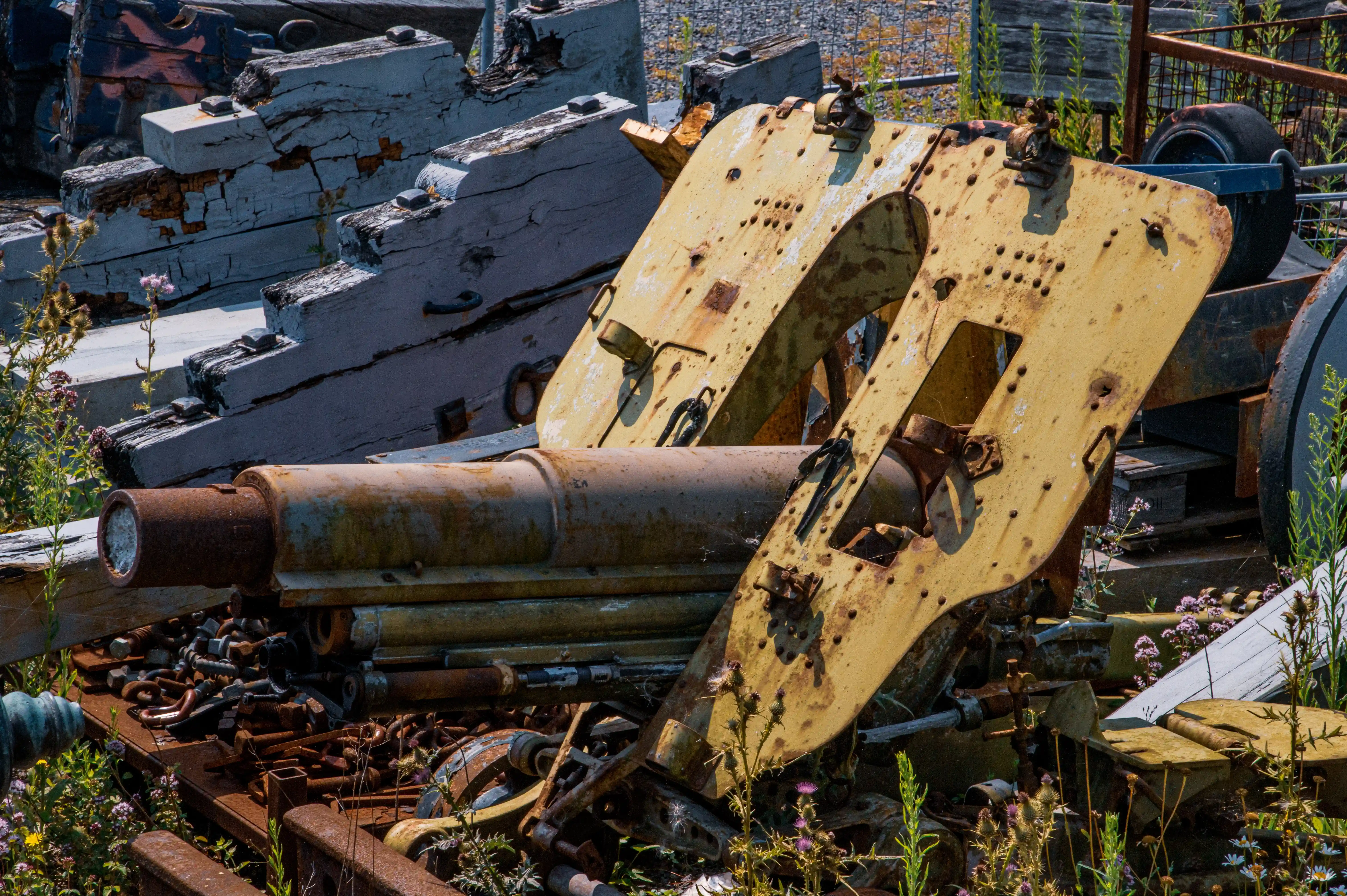 The image depicts an old, rusted piece of machinery, likely a cannon, situated in an outdoor setting with various other rusted metal objects and wooden planks. The machinery is heavily corroded, indicating it has been abandoned for a significant period. The surrounding area is overgrown with plants and wildflowers, suggesting a state of disrepair and neglect. The scene evokes a sense of history and decay, with the machinery possibly being a relic from a past era.
