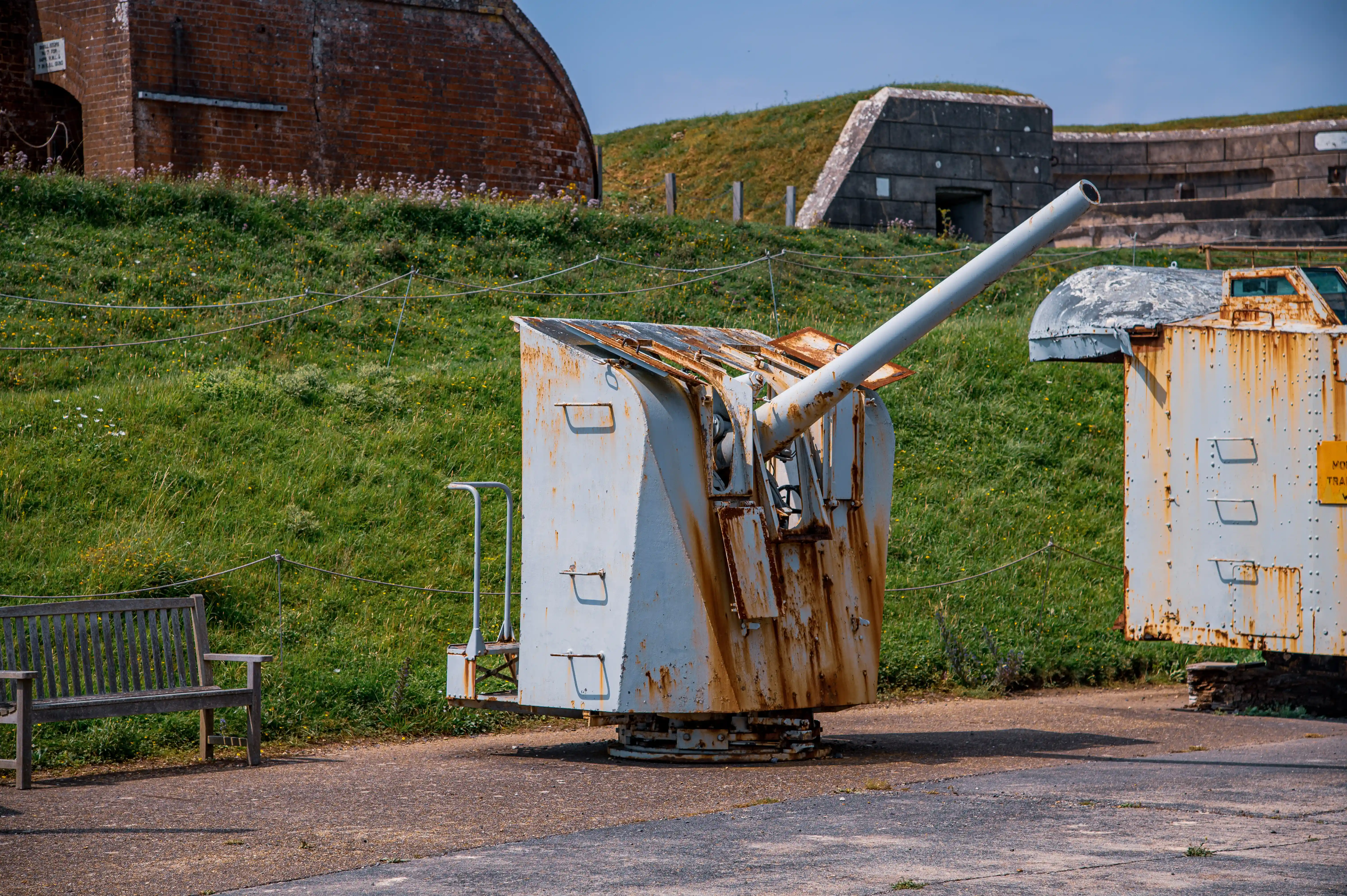 The image depicts an outdoor scene featuring two rusted, military-style structures that appear to be gun turrets or similar artillery equipment. These structures are situated on a grassy hill, with one turret in the foreground and another slightly behind it. The turrets are surrounded by a low barbed wire fence, and there are benches in front of the structures. In the background, there are brick buildings and a concrete bunker, suggesting that this fort might be part of a historical or military site.