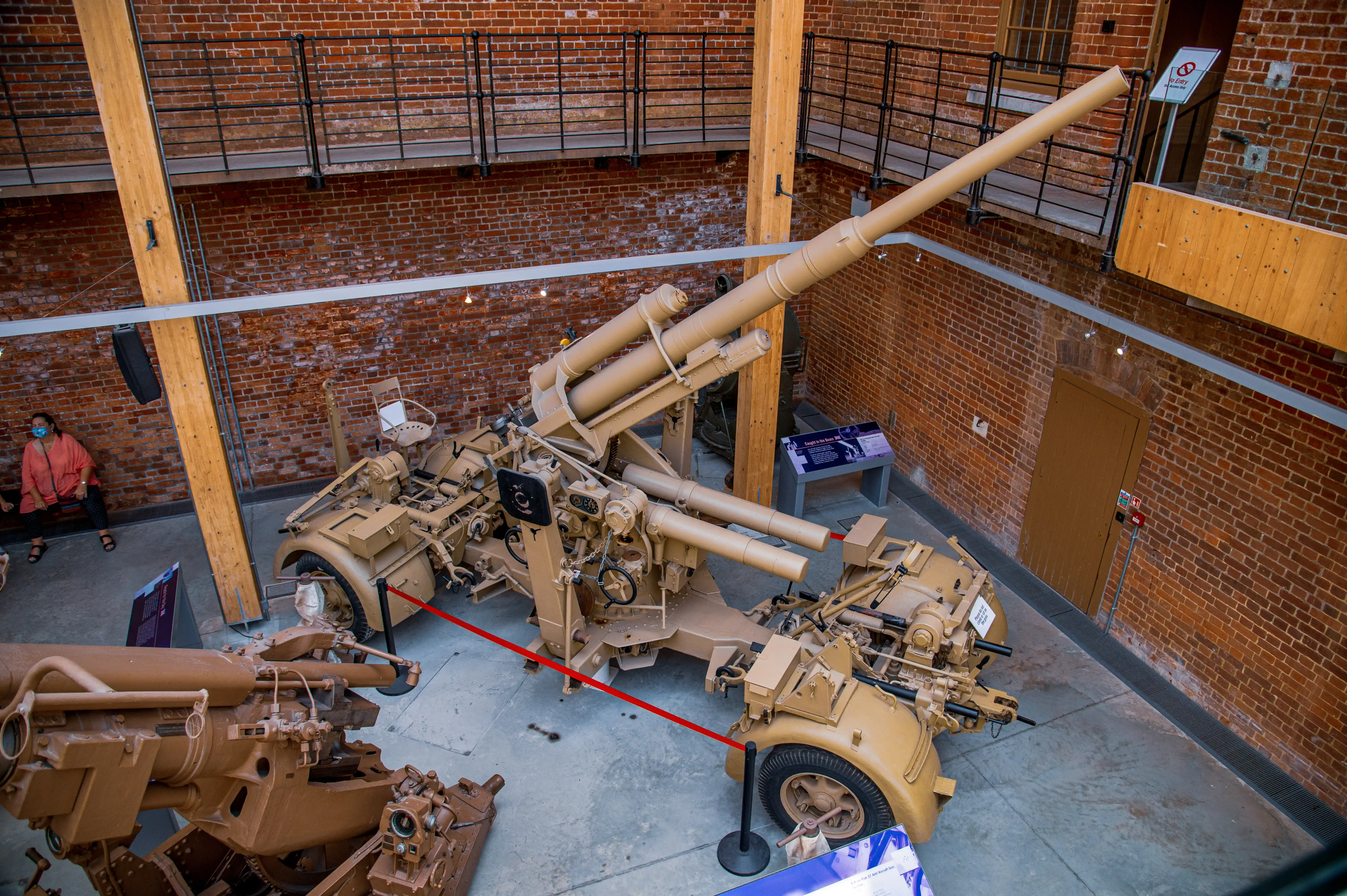 The image depicts a museum exhibit featuring various pieces of military artillery. The display includes several large cannons and artillery pieces, which are cordoned off with red ropes to prevent visitors from touching them. The setting appears to be indoors, with brick walls and wooden structural elements. There is a person in the background, possibly a visitor or a staff member, and informational placards are placed near the exhibits for educational purposes.