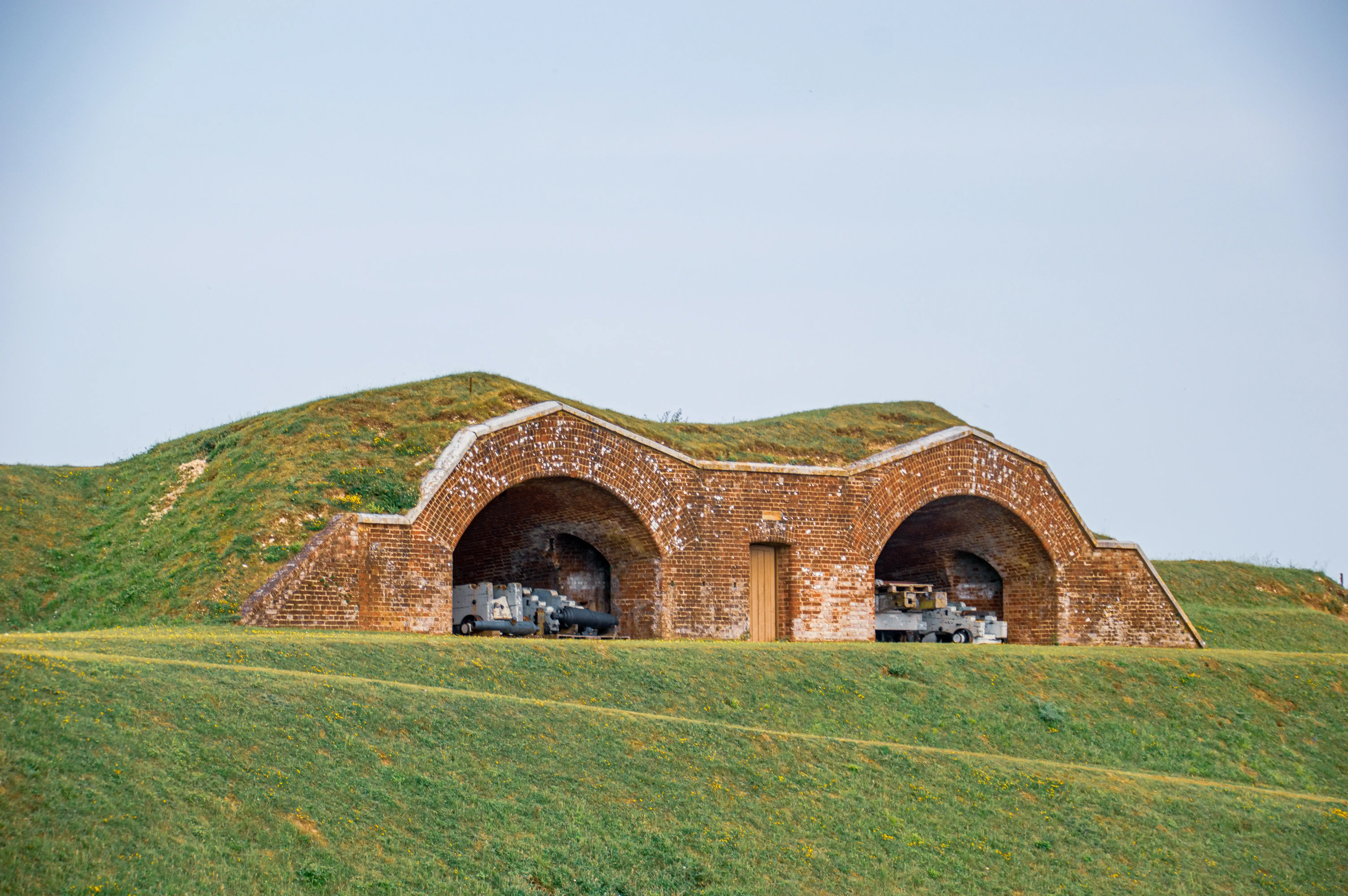 The image depicts a historical fortification structure made of brick with two large arched openings. The structure is partially embedded into a grassy hill, suggesting it may be part of an old fort or defensive wall. Inside the arches, there are what appear to be old cannons or artillery pieces, indicating the site's military significance. The surrounding area is grassy with a clear sky above, giving the scene a peaceful yet historical ambiance.