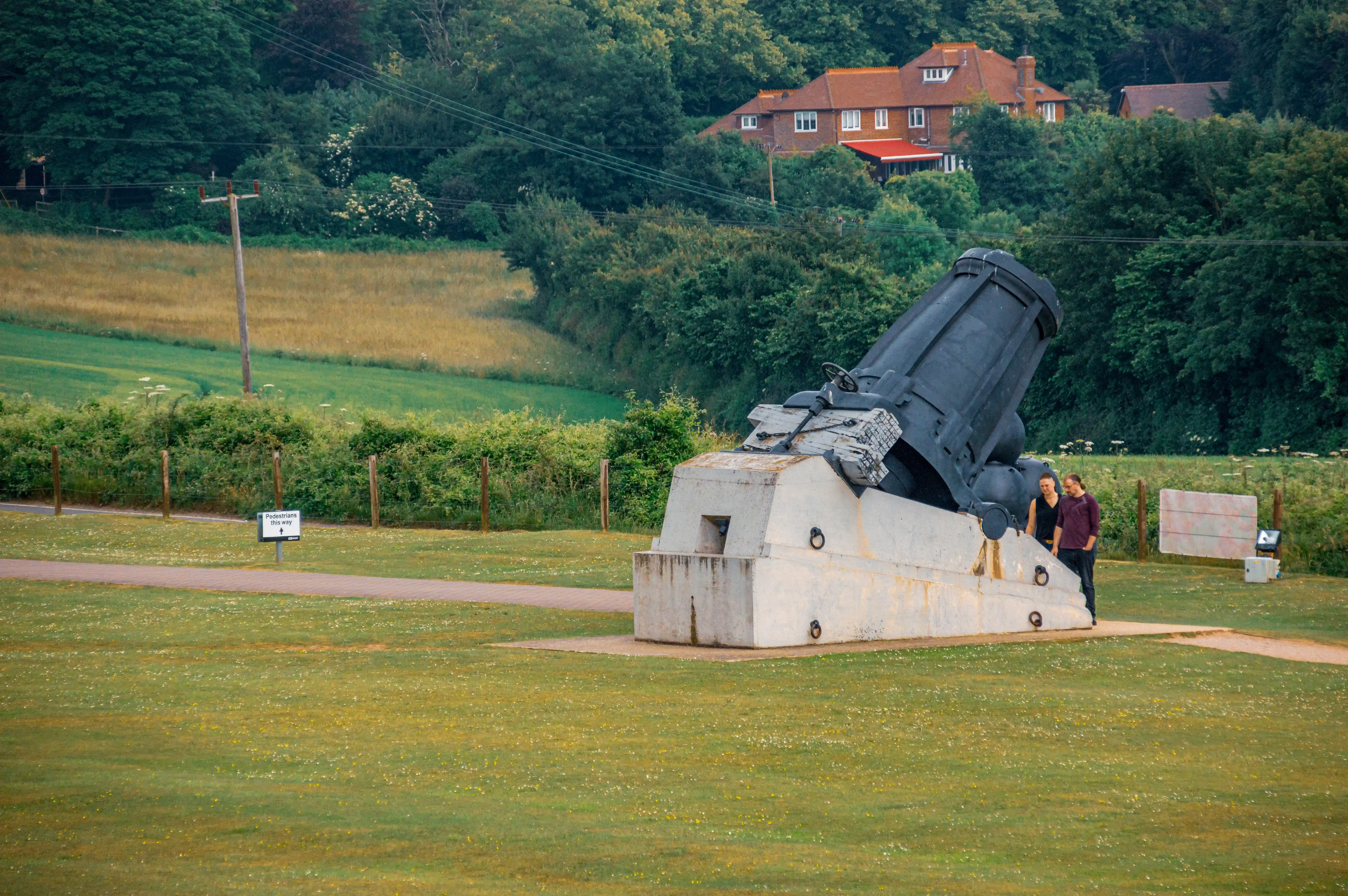 The image depicts a large, historical military artillery piece mounted on a concrete platform in an open, grassy area. Two people are standing next to the artillery, providing a sense of scale. The background features a rural landscape with houses, trees, and fields. There is a sign nearby indicating a pedestrian crossing. The overall setting appears to be a historical site or museum, likely dedicated to military history.