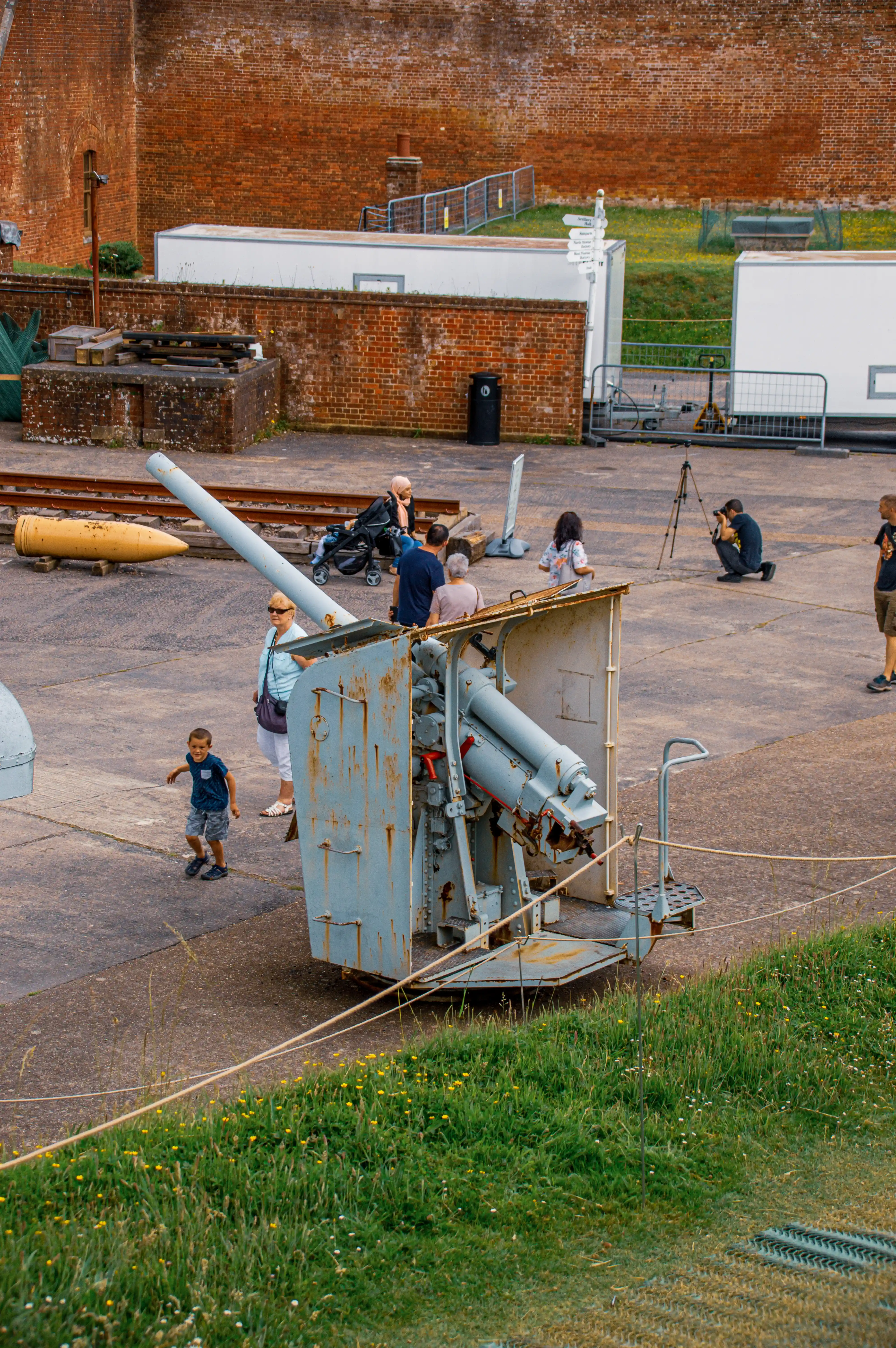 The image shows a group of people, including children and adults, gathered around historical military artillery pieces displayed in an outdoor museum setting. The artifacts include large guns and bombs placed on a concrete surface near a brick wall. The scene is set in a grassy area with some scattered dandelions and a few structures in the background. Some visitors are closely examining the exhibits, while one person is taking photographs. The overall atmosphere suggests an educational or tourist visit to a historical site.