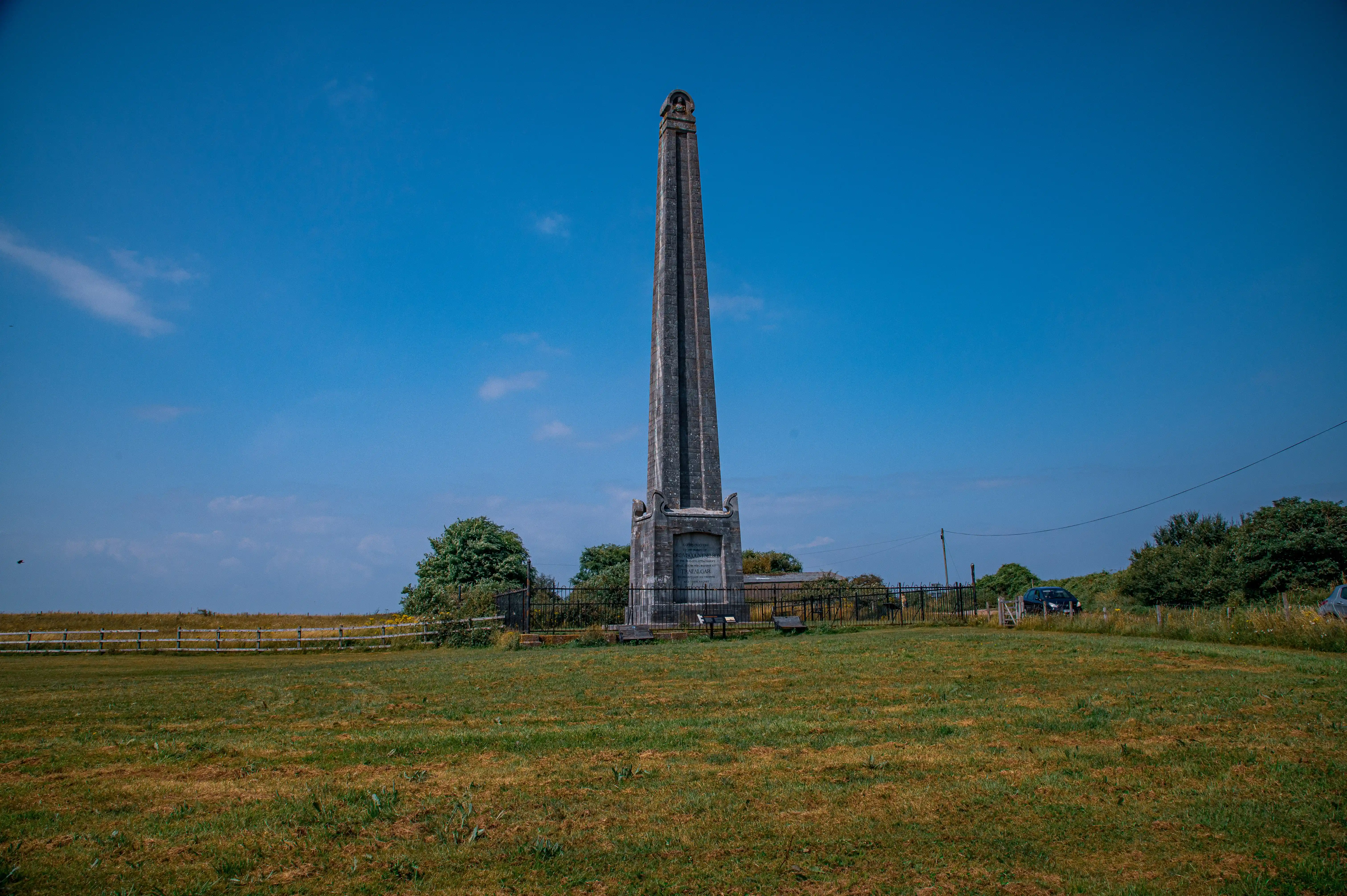 The image depicts a tall, stone obelisk monument situated in an open, grassy field under a clear blue sky. The monument is surrounded by a low fence and is located near a road with a few vehicles parked nearby. The area around the monument is relatively flat and features some trees and shrubs. The monument appears to be well-maintained and is likely of historical or commemorative significance.