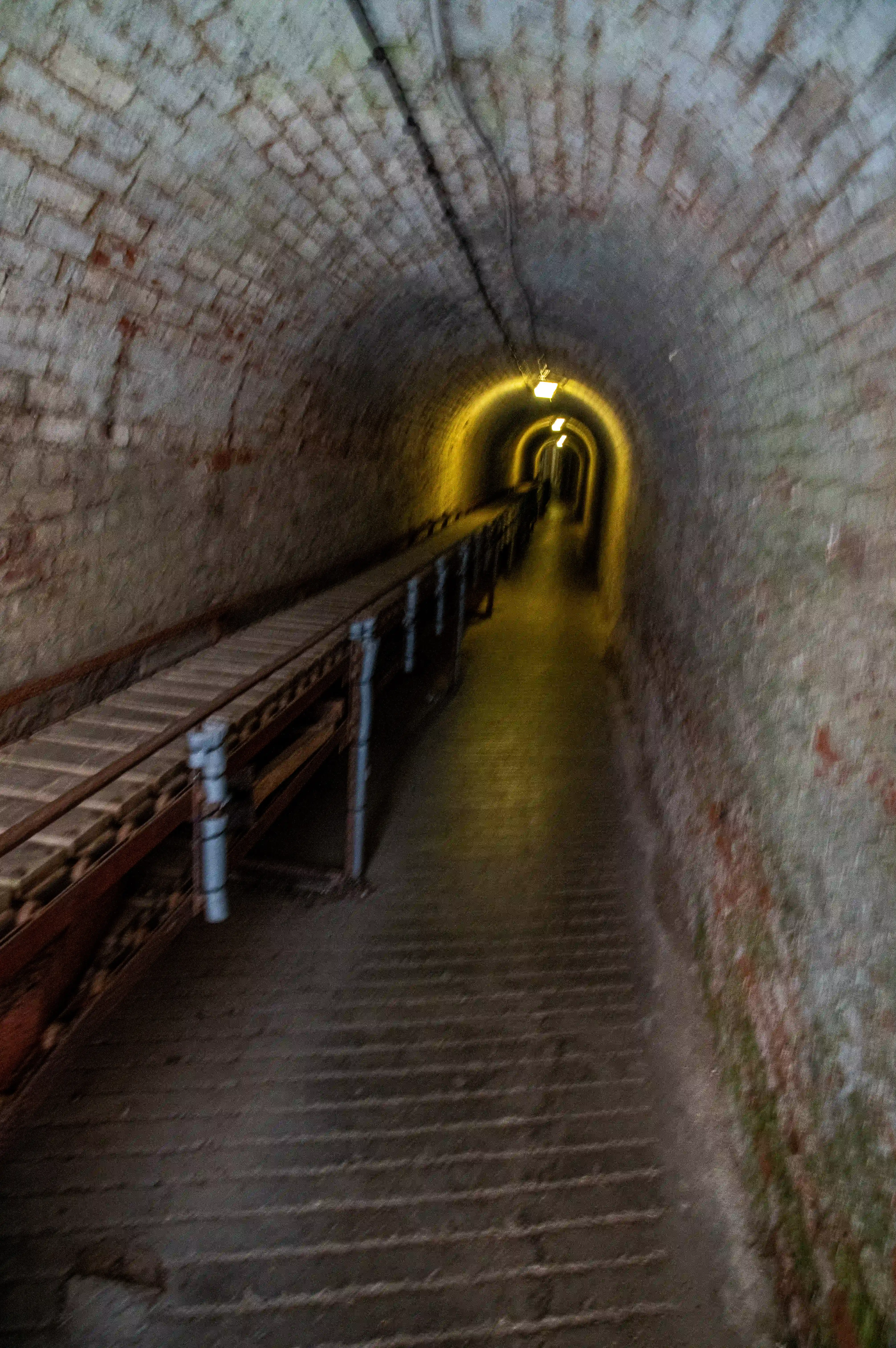 The image depicts a dimly lit, narrow tunnel with a brick floor and walls. The tunnel is illuminated by a series of lights at the far end, creating a warm glow. There are rail tracks running along the left side of the tunnel, suggesting it might be used for a small train or carts. The walls appear aged and weathered, with some patches of discoloration and wear. The perspective of the image gives a sense of depth and distance, leading the viewer's eye towards the light at the end of the tunnel.