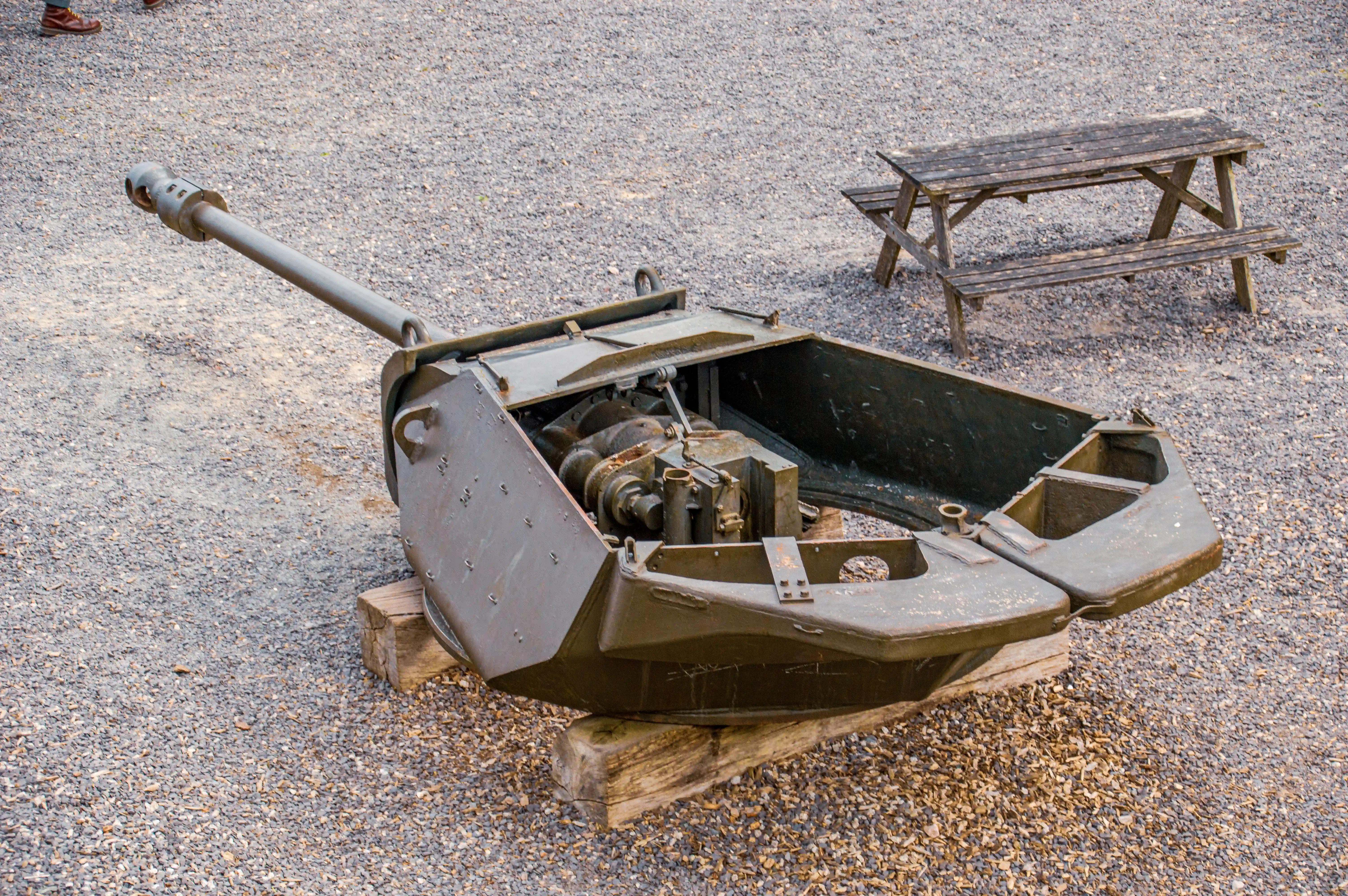 The image depicts an old, rusted, and seemingly abandoned piece of machinery, possibly a tank turret or similar military equipment, placed on a wooden platform. The machinery is open, revealing its internal components. The setting appears to be an outdoor area with gravel ground and a wooden bench in the background.