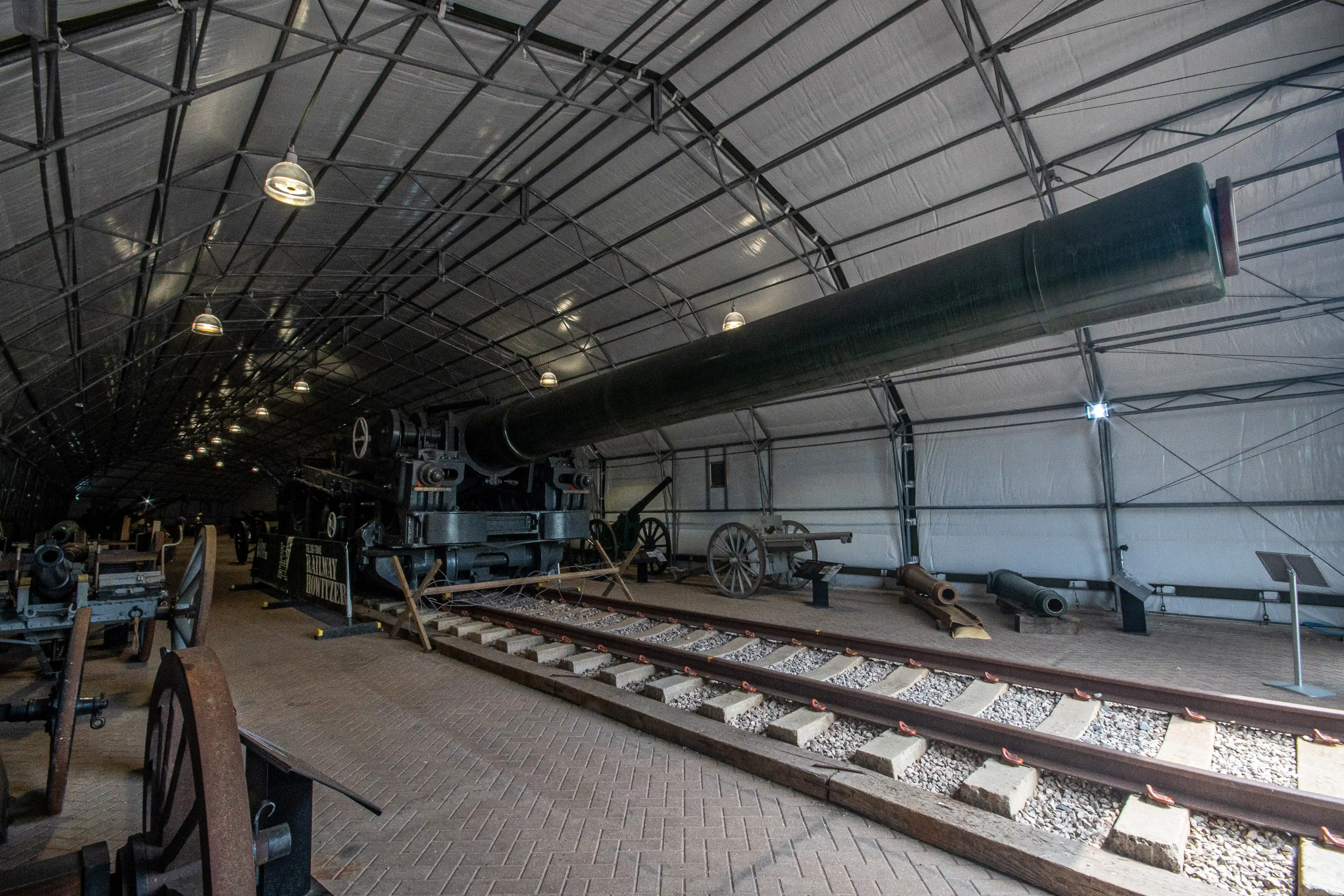 The image depicts an indoor display of historical artillery and railway equipment. The setting appears to be a museum or exhibition hall with a high, tent-like ceiling supported by a metal framework. The display includes a large, old-fashioned cannon mounted on a railway track, several smaller cannons, and various other historical military artifacts. Informational plaques are placed near the exhibits, providing context and details about the items on display.