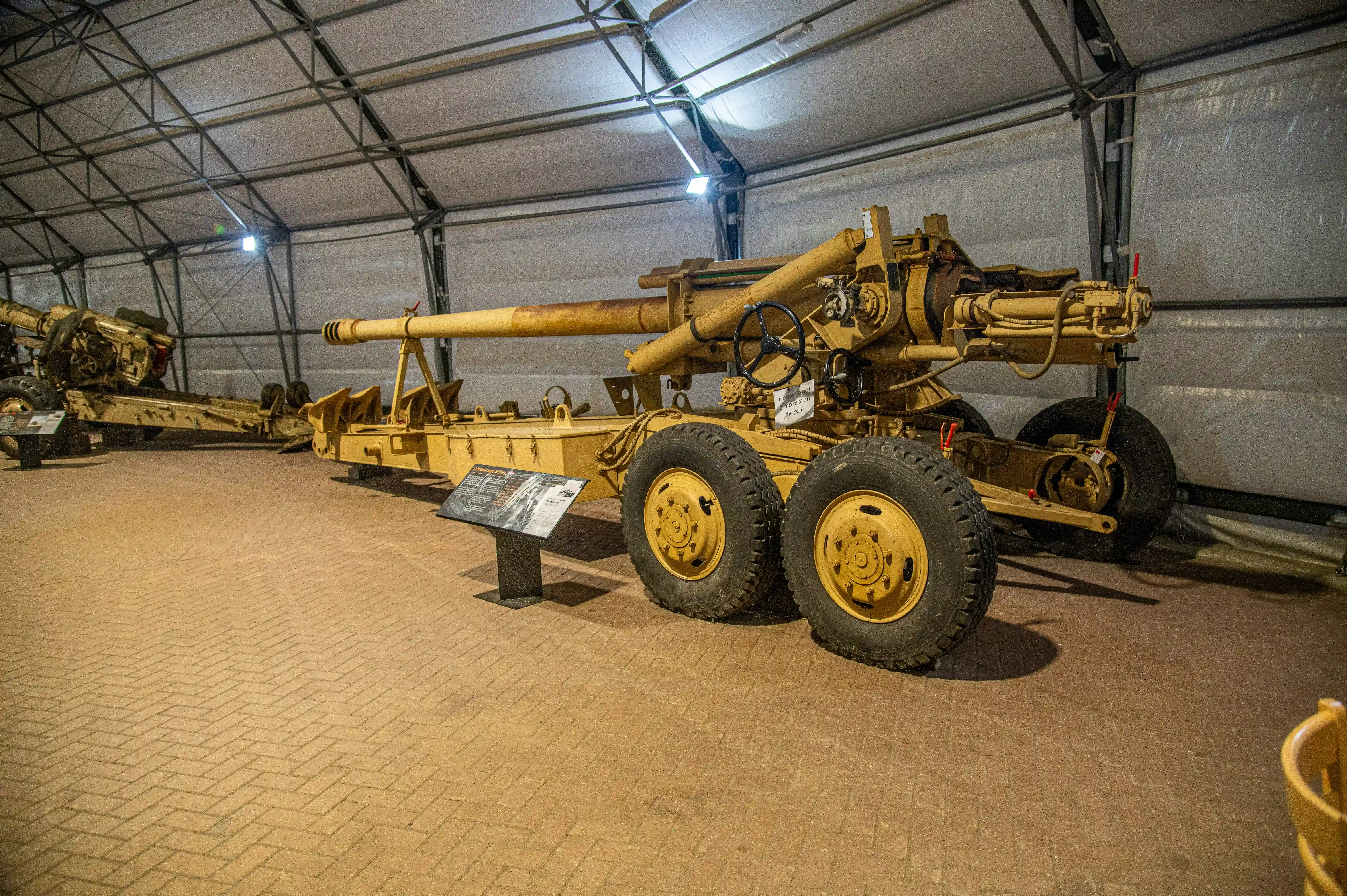 The image shows a military artillery piece displayed indoors, likely in a museum setting. The artillery is mounted on a wheeled carriage and is positioned on a paved floor. There is an informational plaque in front of the artillery, providing details about the exhibit. The setting appears to be a large, well-lit tent or temporary structure with a high ceiling.