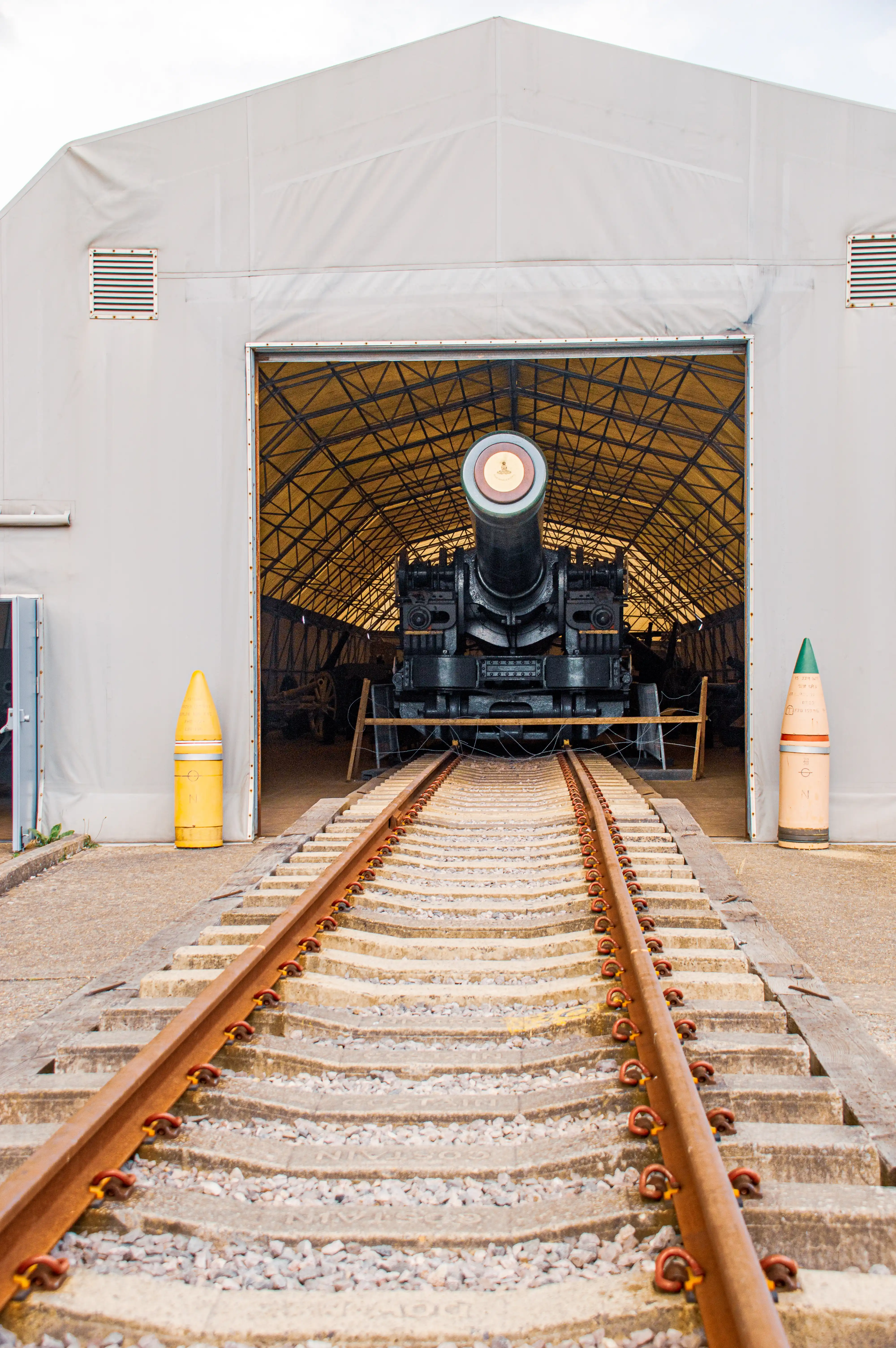The image shows a large cannon mounted on a railway track inside a tent-like structure. The cannon is positioned at the end of the tracks, facing outward. The tent is open in the front, revealing the cannon and part of the interior. There are large projectile shells on either side of the entrance.