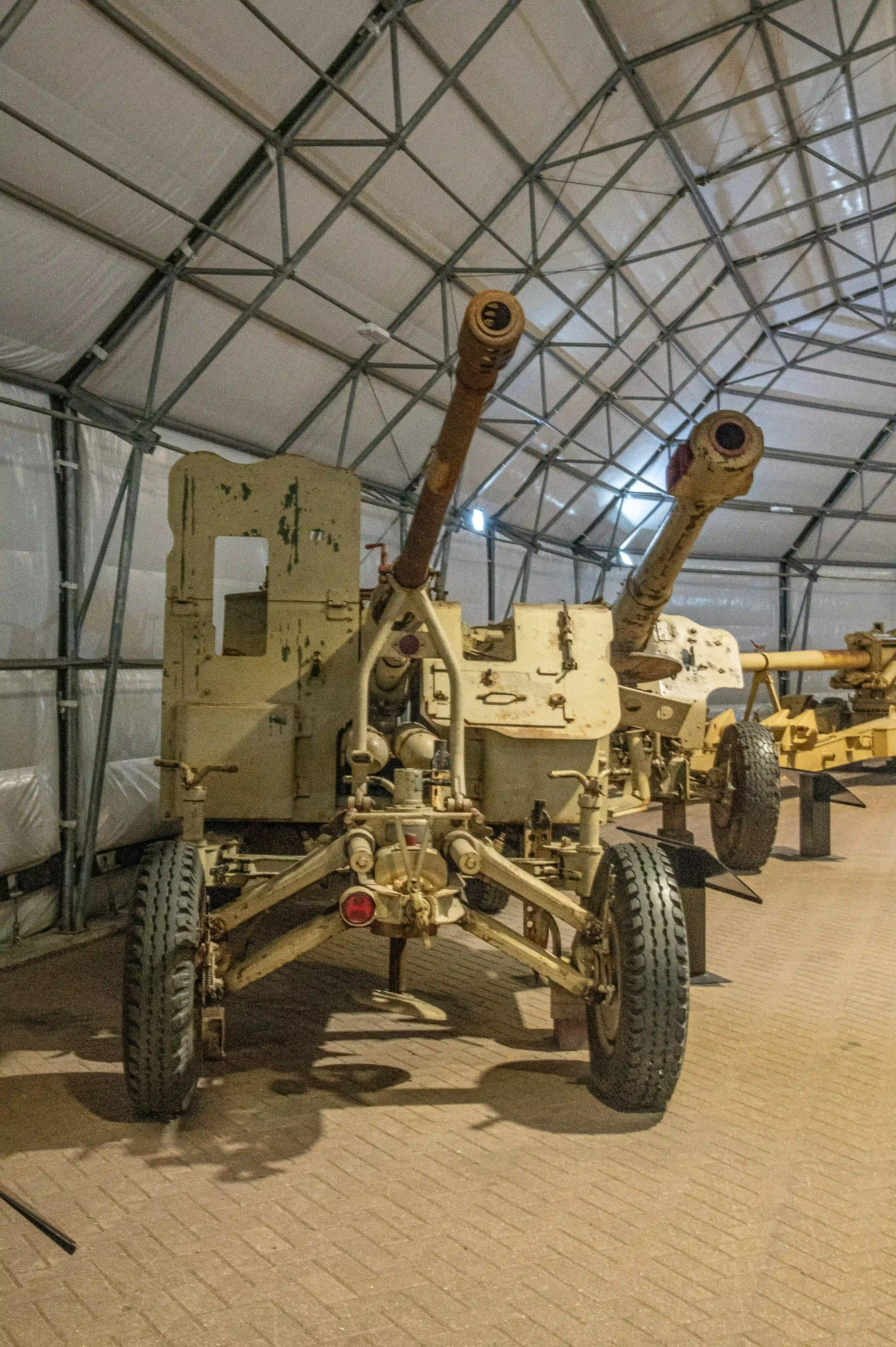 The image shows a collection of military artillery pieces displayed indoors under a large tent-like structure. The artillery pieces are mounted on wheeled carriages and appear to be historical, possibly from World War II or earlier. The setting suggests a museum or an exhibition space dedicated to military history. The artillery pieces are arranged in a line, and the environment is well-lit, highlighting the details of the equipment.