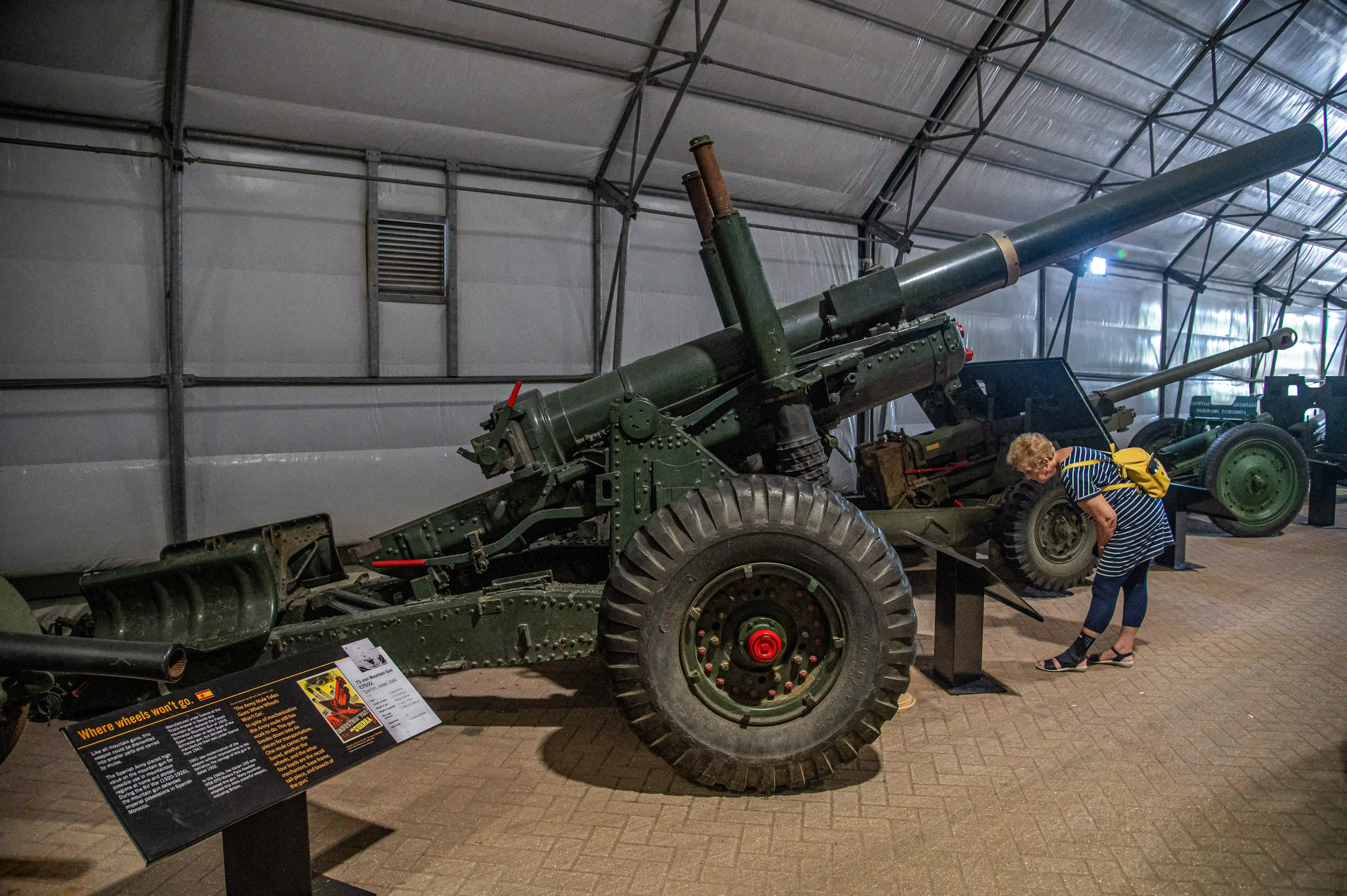 The image shows a museum exhibit featuring large artillery pieces. A person is closely examining one of the cannons, which is mounted on wheels and has a large barrel. Informational placards are present, providing details about the artillery. The setting appears to be an indoor exhibit space with a high ceiling and a concrete floor.