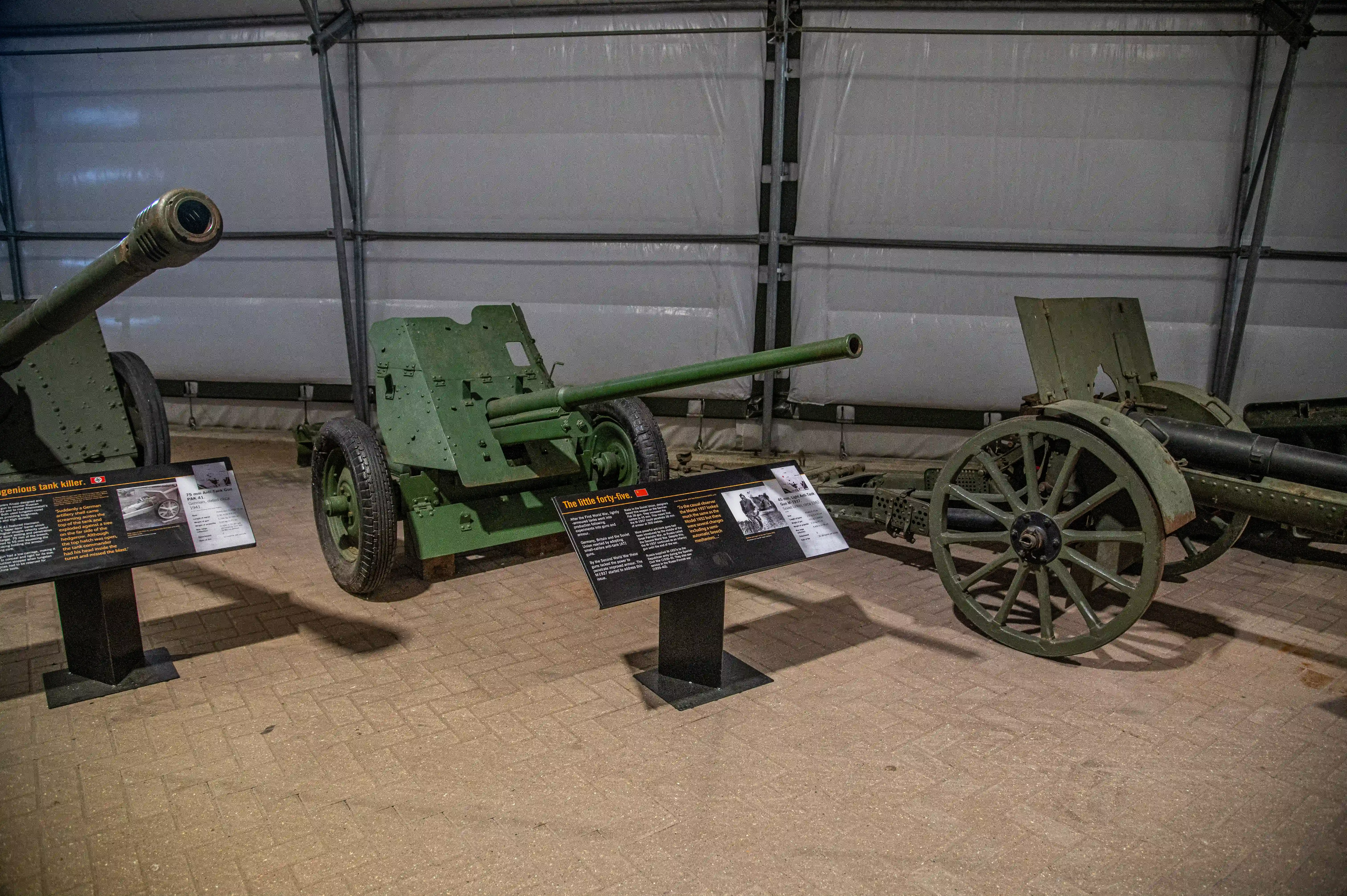 The image depicts an indoor display of historical artillery pieces in a museum setting. There are three cannons on display, each with an accompanying informational placard. The cannons are positioned on wooden platforms, and the background features a large, gray, industrial-style curtain. The placards provide detailed information about each cannon, including their historical significance and specifications.