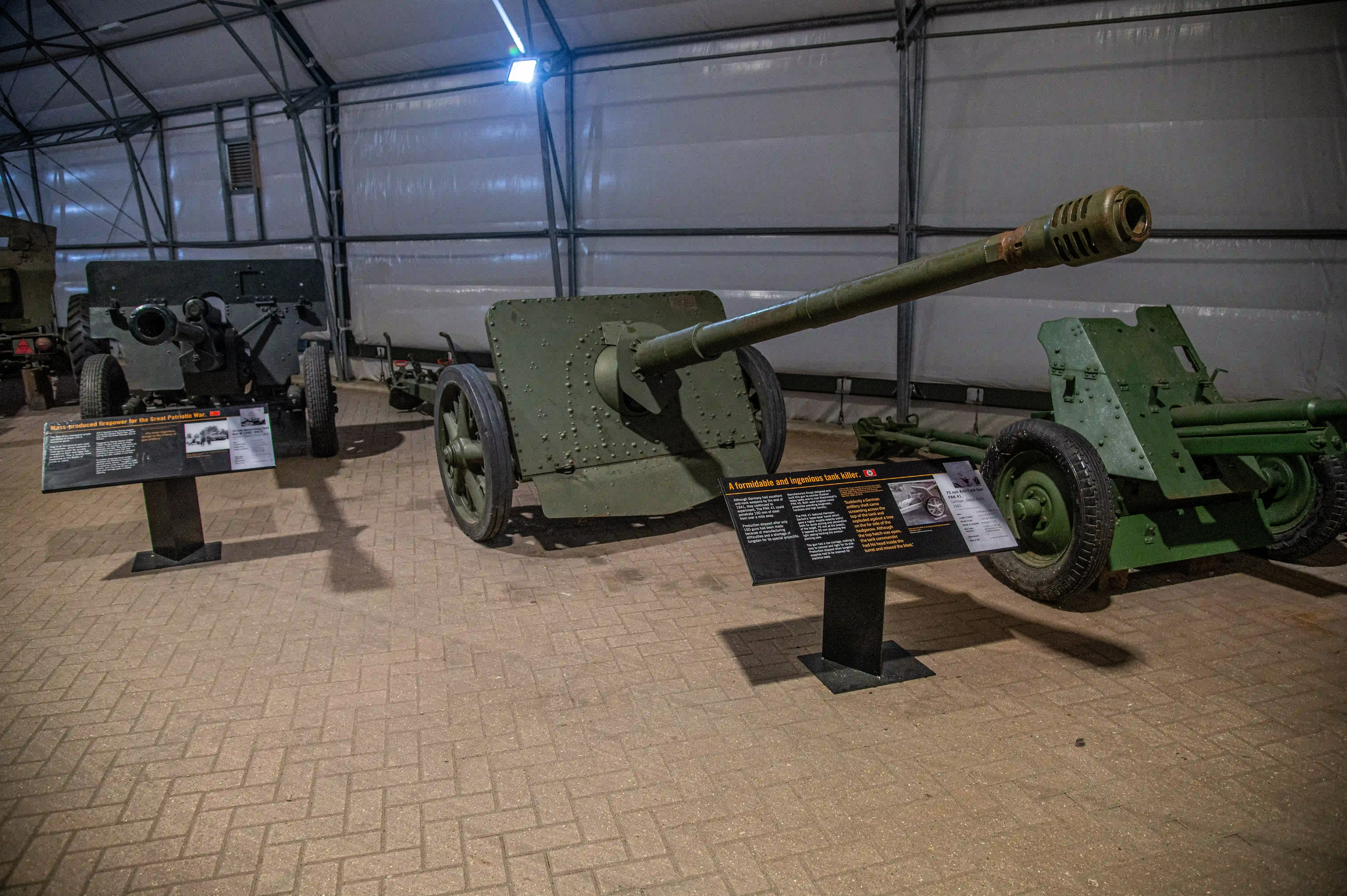 The image shows an indoor display of historical artillery pieces, likely from a museum. The exhibit includes a large green cannon in the foreground with an informational placard in front of it, detailing its historical significance. In the background, another artillery piece is visible, also with an accompanying placard. The setting appears to be a well-lit, spacious area with a brick floor, suitable for showcasing these military artifacts.