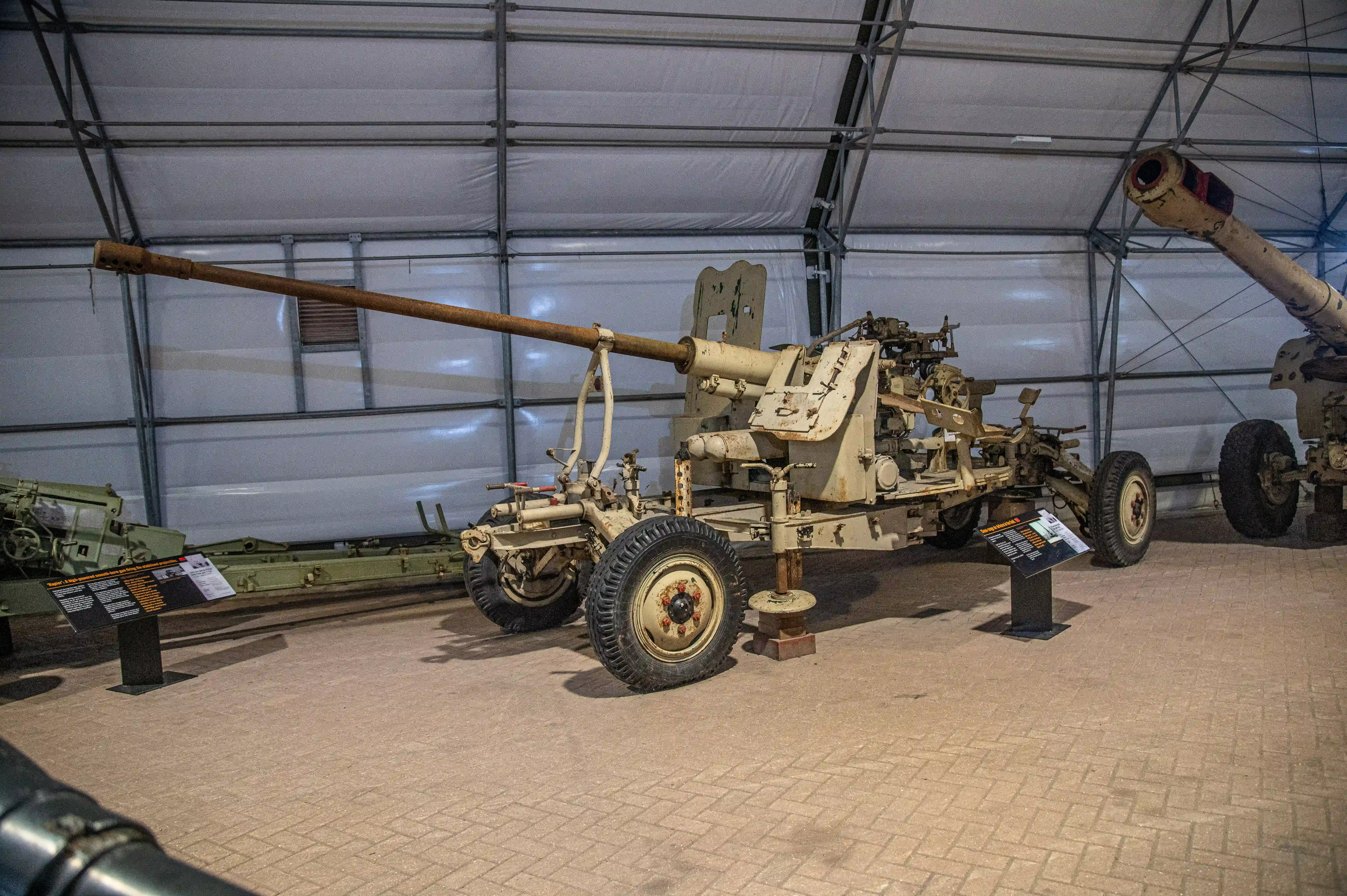 The image depicts a collection of historical artillery pieces displayed in a museum-like setting. The main focus is on a large, wheeled artillery gun in the center, which appears to be from the mid-20th century. Surrounding it are other artillery pieces, including a smaller field gun on the left and a large naval or coastal gun on the right. Each piece is mounted on stands with informational plaques in front of them, providing details about their history and specifications. The setting is indoors, with a large, white tent-like structure providing a backdrop..