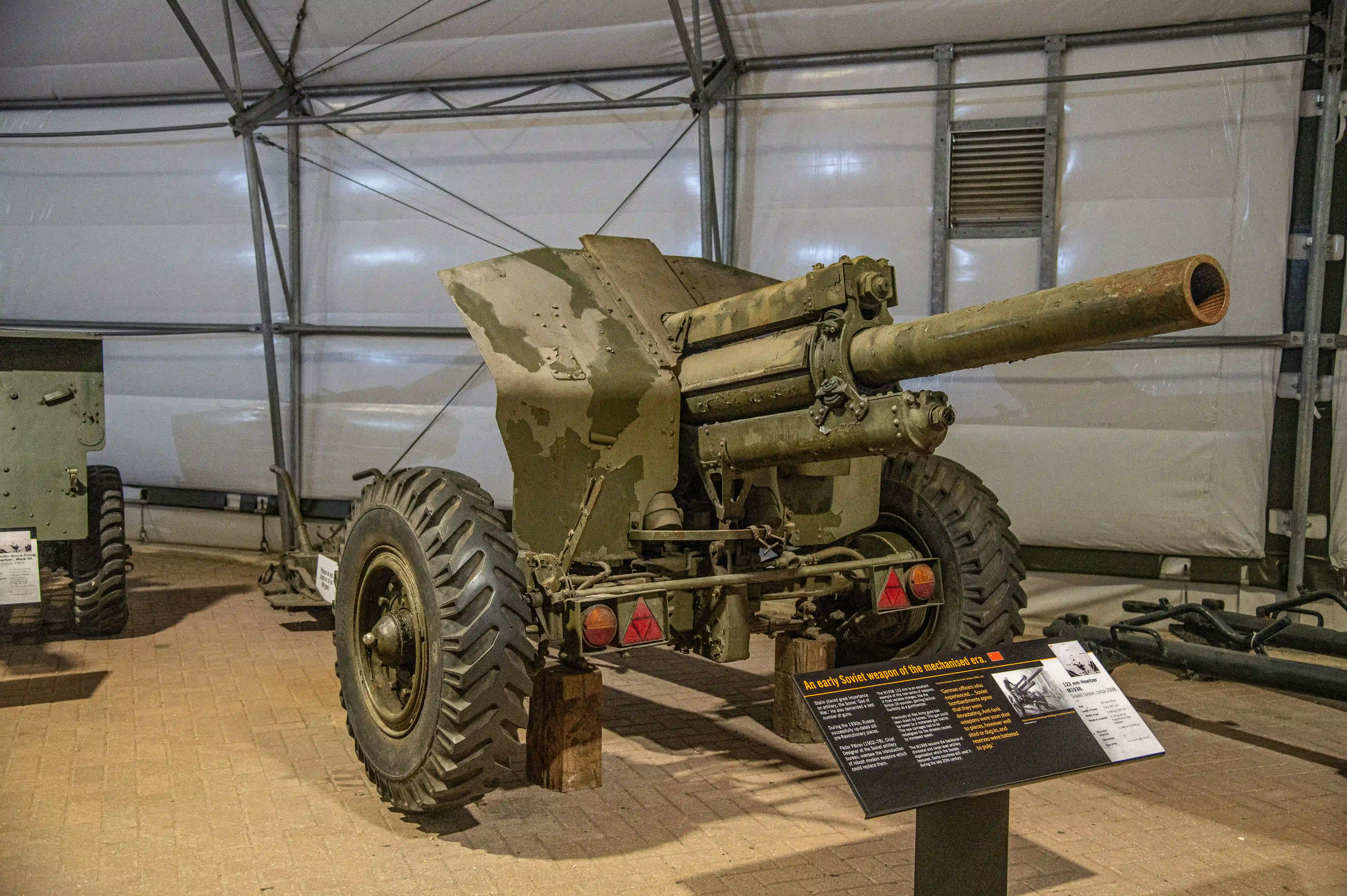The image depicts an artillery piece displayed in a museum setting. The artillery is mounted on large wheels and is positioned inside a tent-like structure. There is an informational placard in front of the artillery, providing details about its historical significance, likely indicating it is an early Soviet weapon from the mechanized era. The surroundings include other military equipment and exhibits.