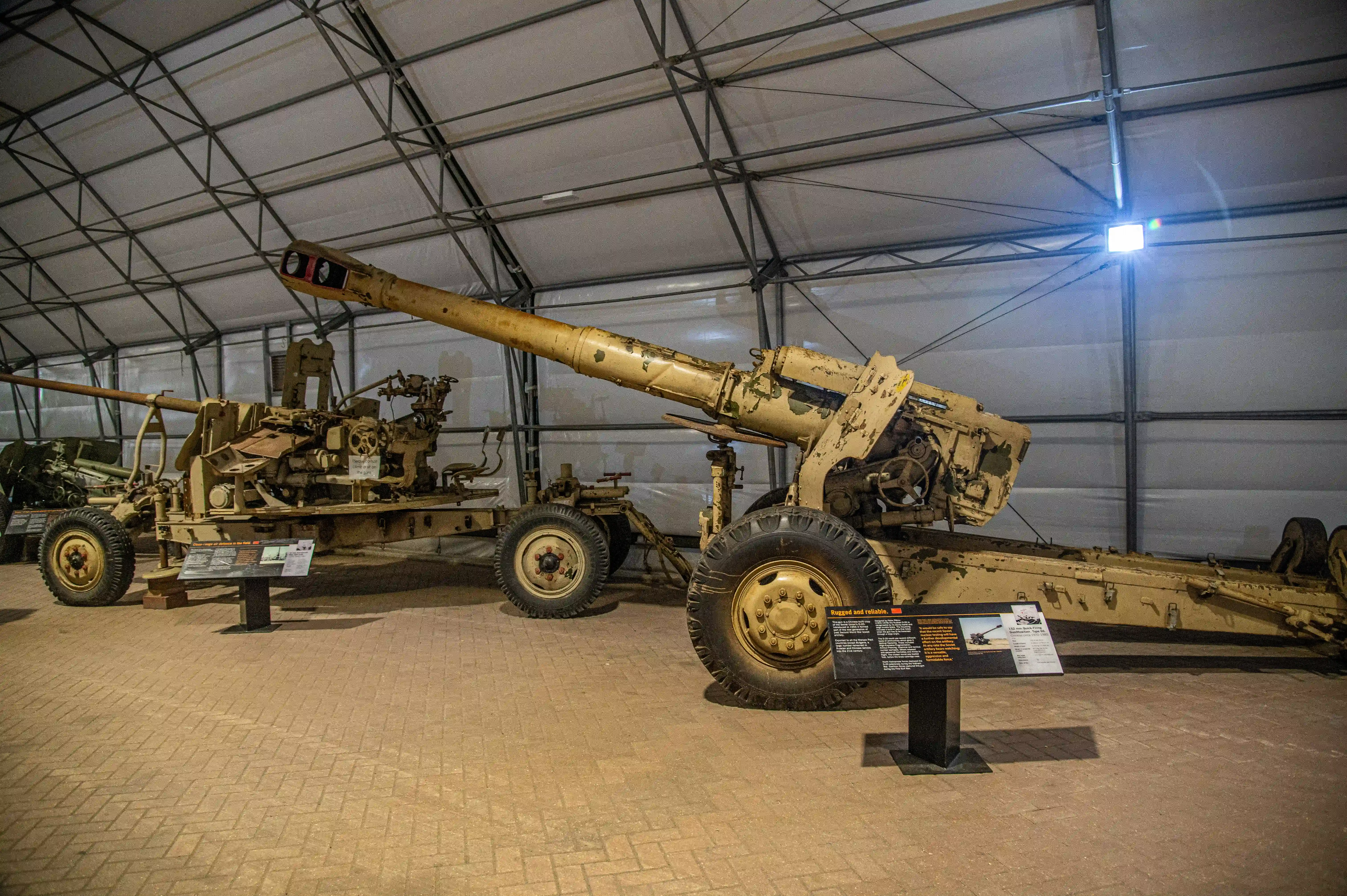 The image shows a military artillery piece displayed in a museum setting. The artillery is mounted on a wheeled carriage and is positioned inside a large, tent-like structure. There are informational plaques in front of and beside the artillery, providing details about the exhibit. The surroundings are clean and well-lit, indicating a well-maintained display area.