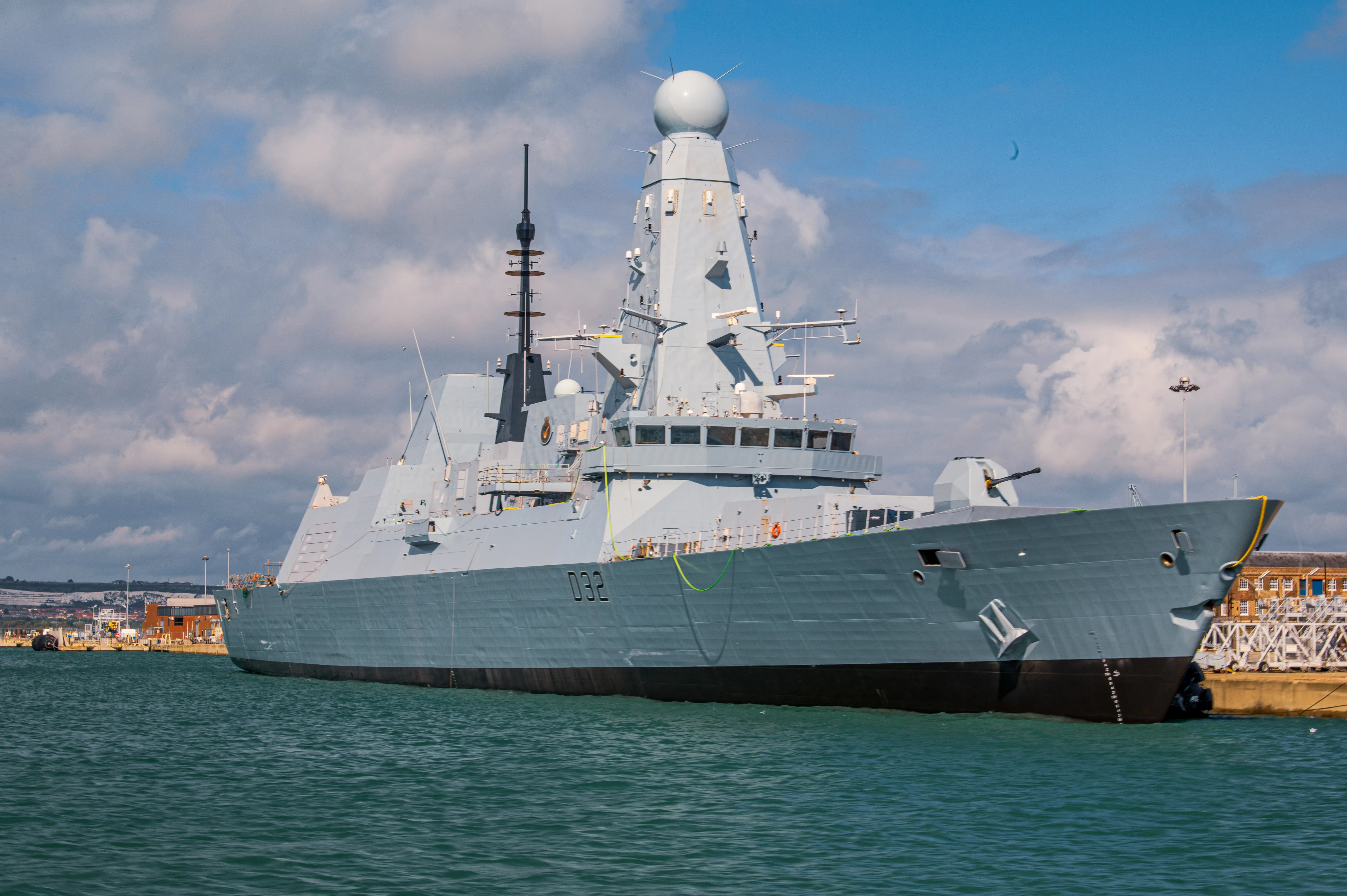 The image shows a large naval ship docked at a port. The ship is grey with a distinctive angular design and various antennas and equipment on its deck. The number 032 is visible on the side of the ship. The background includes a cityscape with buildings and a partly cloudy sky. The water in the foreground is calm and greenish in color.