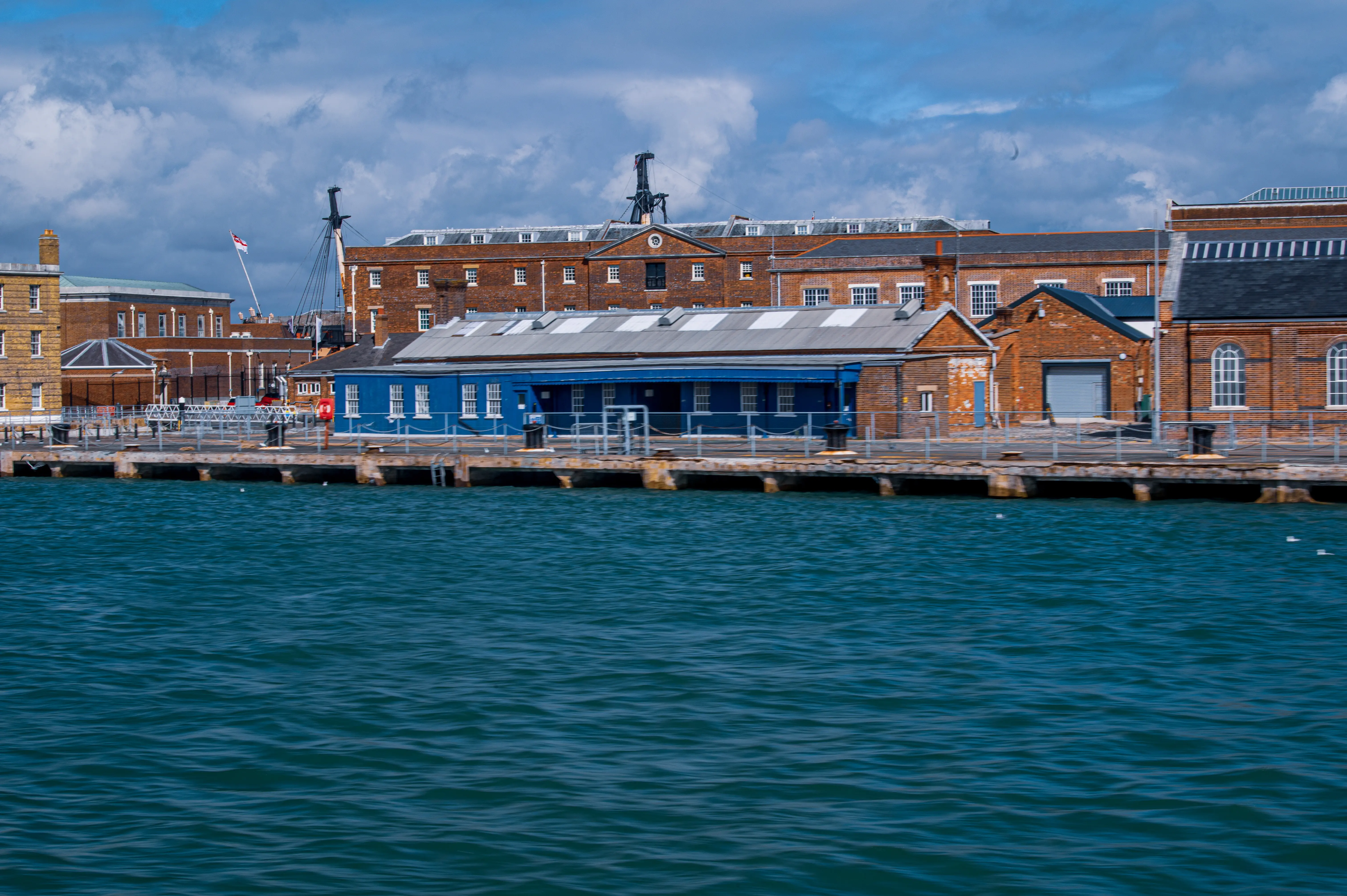 The image shows a waterfront scene featuring a large, historic brick building with multiple sections and architectural details. The building has a prominent clock and flagpole, and there is a smaller blue structure in front of it. The area appears to be a dock or pier, with a calm body of water in the foreground and a partly cloudy sky above.