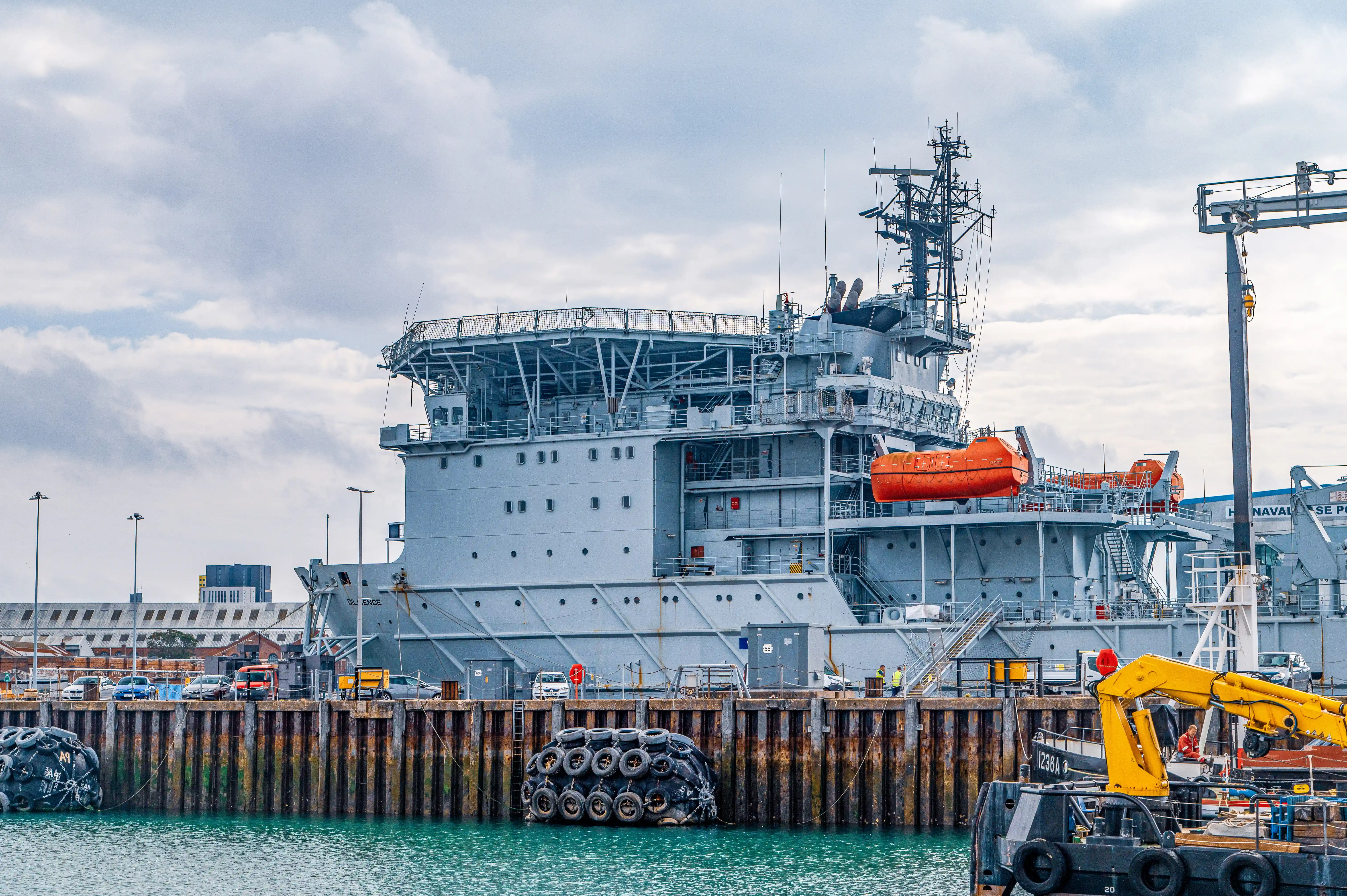 The image depicts a large naval ship docked at a port. The ship is equipped with various antennas and communication equipment on its mast. There are orange lifeboats visible on the side of the ship, and several safety and operational features are evident. The ship is docked next to a wooden quay, where various equipment and vehicles, including a yellow excavator and a tugboat, are present. The background shows additional port infrastructure and other ships.