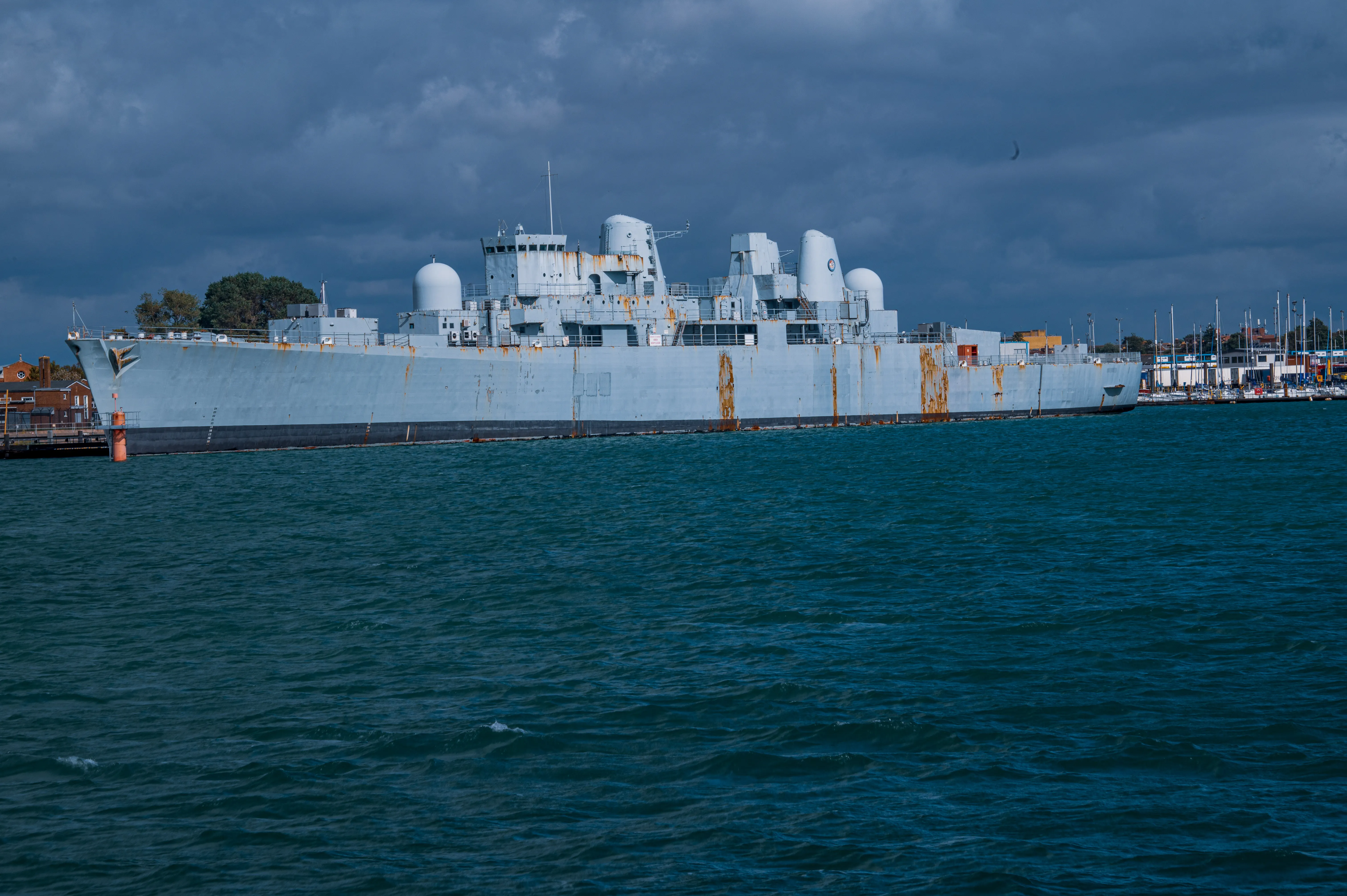 The image depicts a large naval ship docked at a port. The ship has a grey hull with some rust marks and features several radar domes and antennas on its deck. The water in the foreground is a deep blue-green, and the sky above is overcast with dark clouds. The ship appears to be a military vessel, possibly a destroyer or a frigate, given its size and the equipment visible on deck.
