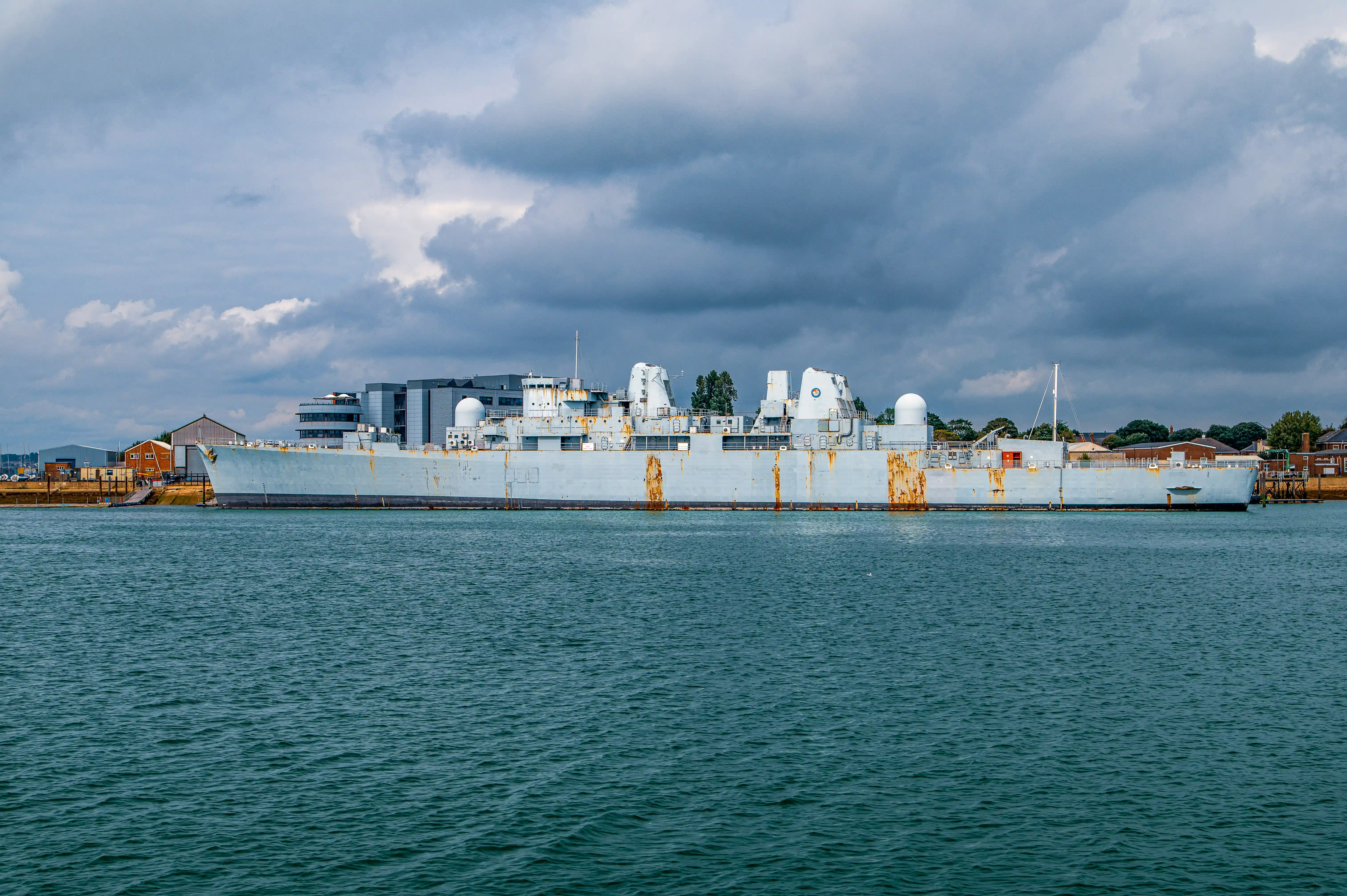 The image depicts a large, weathered naval ship docked at a port. The ship appears to be an aircraft carrier or a similar type of vessel, characterized by its flat deck and various antenna structures. The sky above is overcast with dark clouds, suggesting impending rain. The surrounding area includes industrial buildings and some greenery in the background. The water in the foreground is calm, reflecting the ship and the sky.