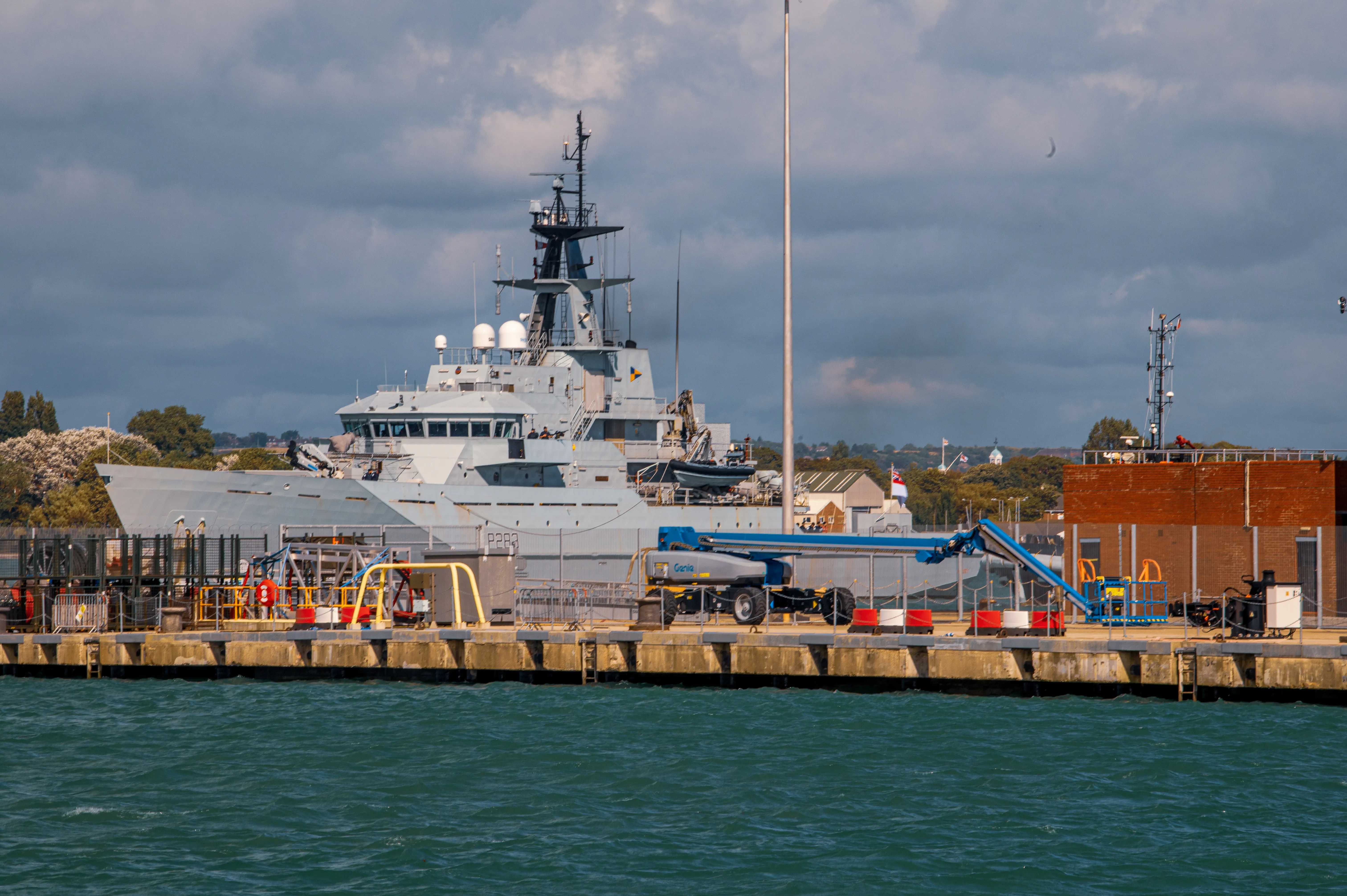 The image shows a large naval vessel docked at a port. The ship is equipped with various antennas and radar equipment, indicating it is likely used for surveillance or command purposes. The dock area has several pieces of equipment and barriers, suggesting ongoing maintenance or preparation activities. The surrounding area includes a mix of industrial and natural elements, with brick buildings and greenery visible in the background. The sky is overcast, contributing to a somewhat muted and serious atmosphere.