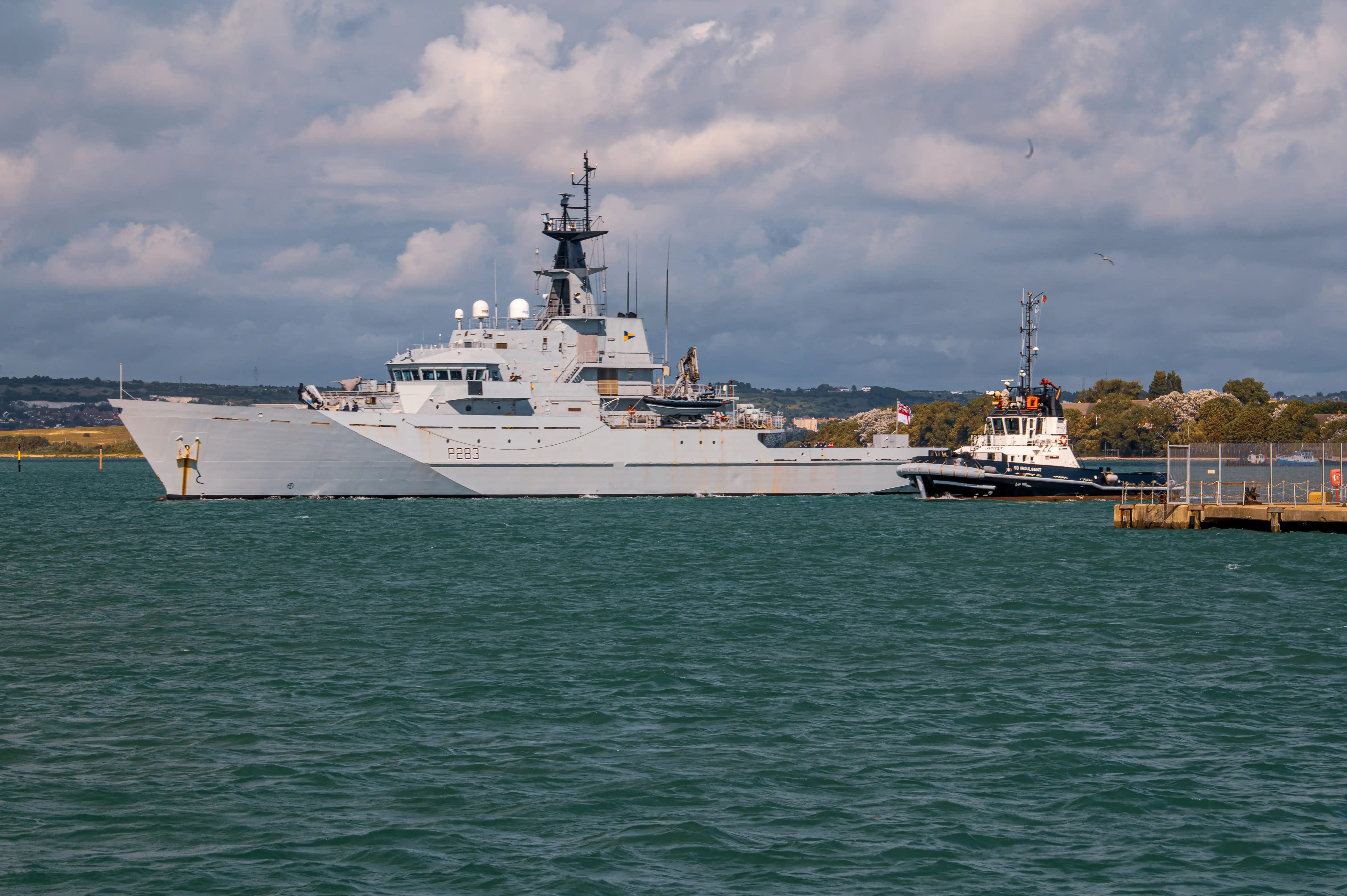 The image depicts a large naval vessel docked at a port. The ship, marked with the identification number P283, is grey and equipped with various antennas and radar equipment on its mast. A smaller tugboat is assisting the naval vessel, which is docked at a concrete pier. The scene is set against a backdrop of a partly cloudy sky and a distant shoreline with trees and hills.