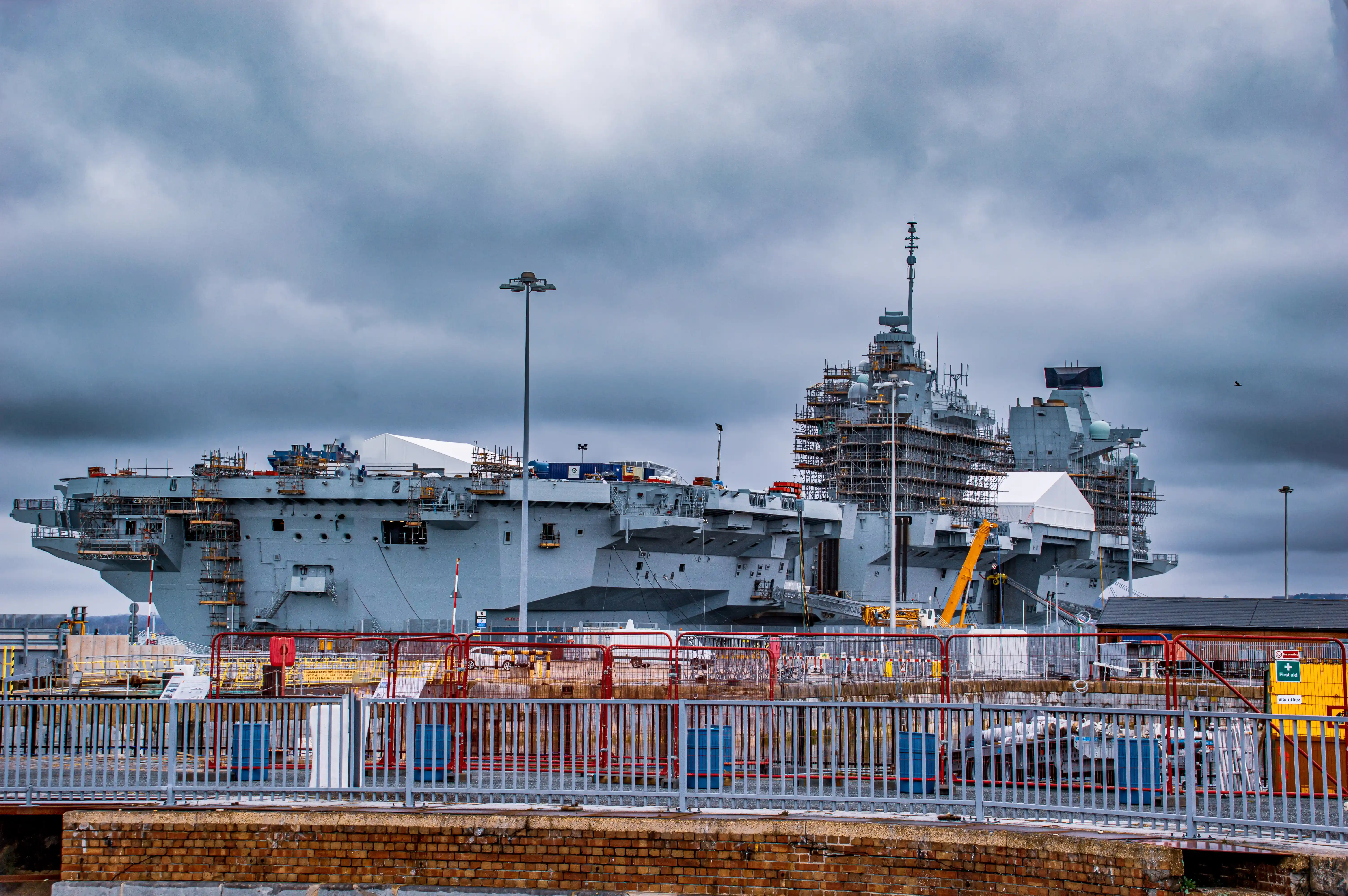 The image shows a large naval ship under construction or maintenance at a dockyard. The ship is surrounded by scaffolding, indicating ongoing work. The sky is overcast, suggesting a cloudy day. The area is secured with fencing and various equipment and materials are visible around the ship. The ship appears to be an aircraft carrier, given its size and structure.